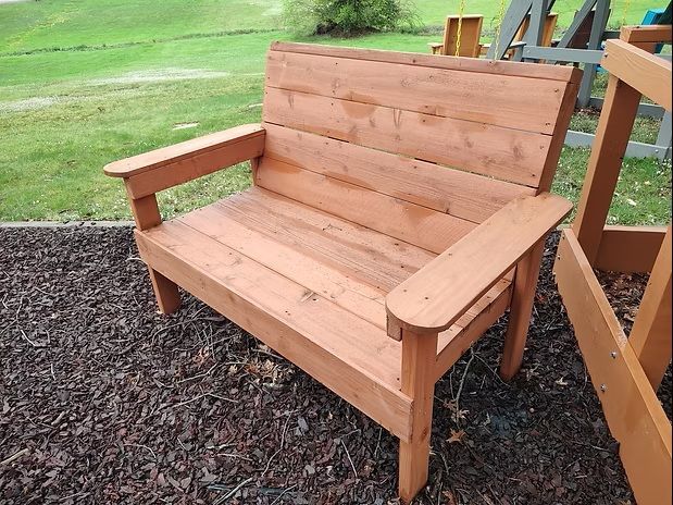 Wooden bench on wood chips, in an outdoor setting. Features armrests and back, appears newly constructed.