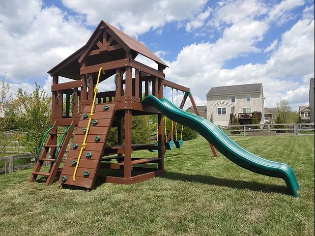 Brown wooden playset with a green slide, climbing wall, and swingset, set on grass.