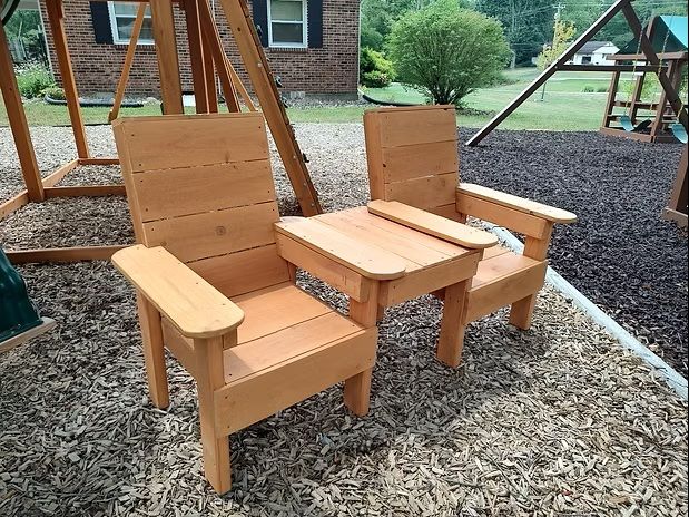 Two wooden outdoor chairs with a connecting side table on mulch, near a swing set.