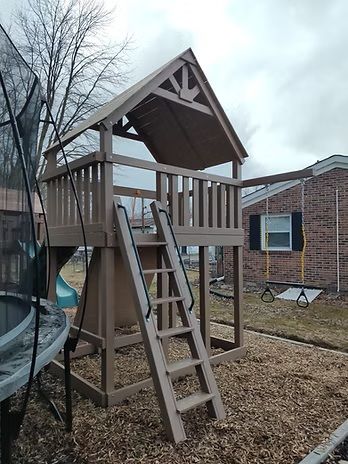 Wooden playset with a house-like structure and a ladder, on wood chips, with a trampoline nearby.