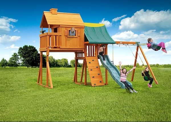Wooden playset with kids: slide, swings, and climbing wall; children playing in a grassy yard.