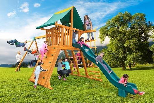 Children playing on a wooden playground with swings, a slide, climbing wall, and rope ladder; sunny day.