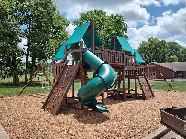 Playground with a green slide, climbing walls, and wooden structure on wood chips, under blue sky.