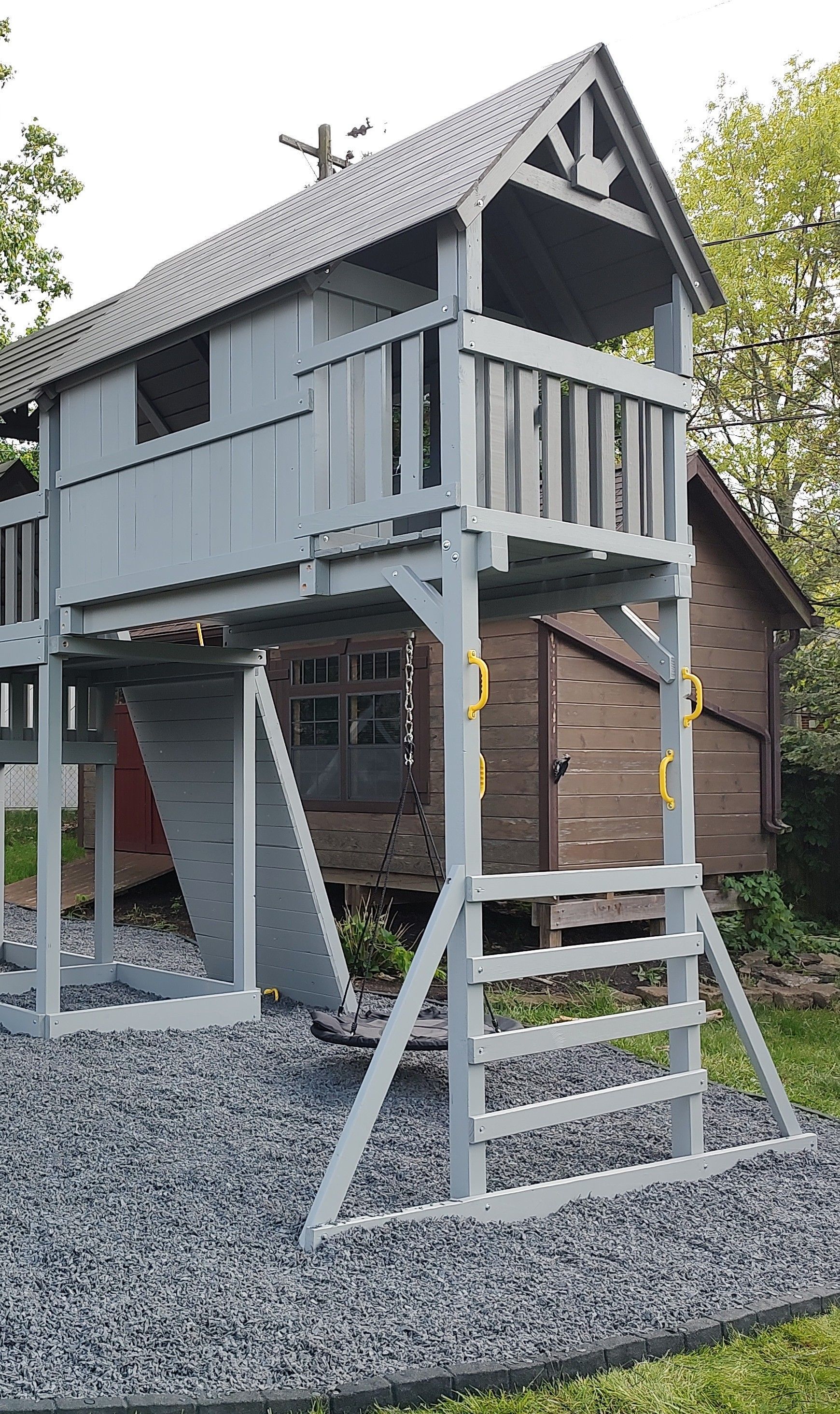 Gray elevated treehouse with wooden stairs and a small balcony on a rubber mulch bed, beside a rustic building
