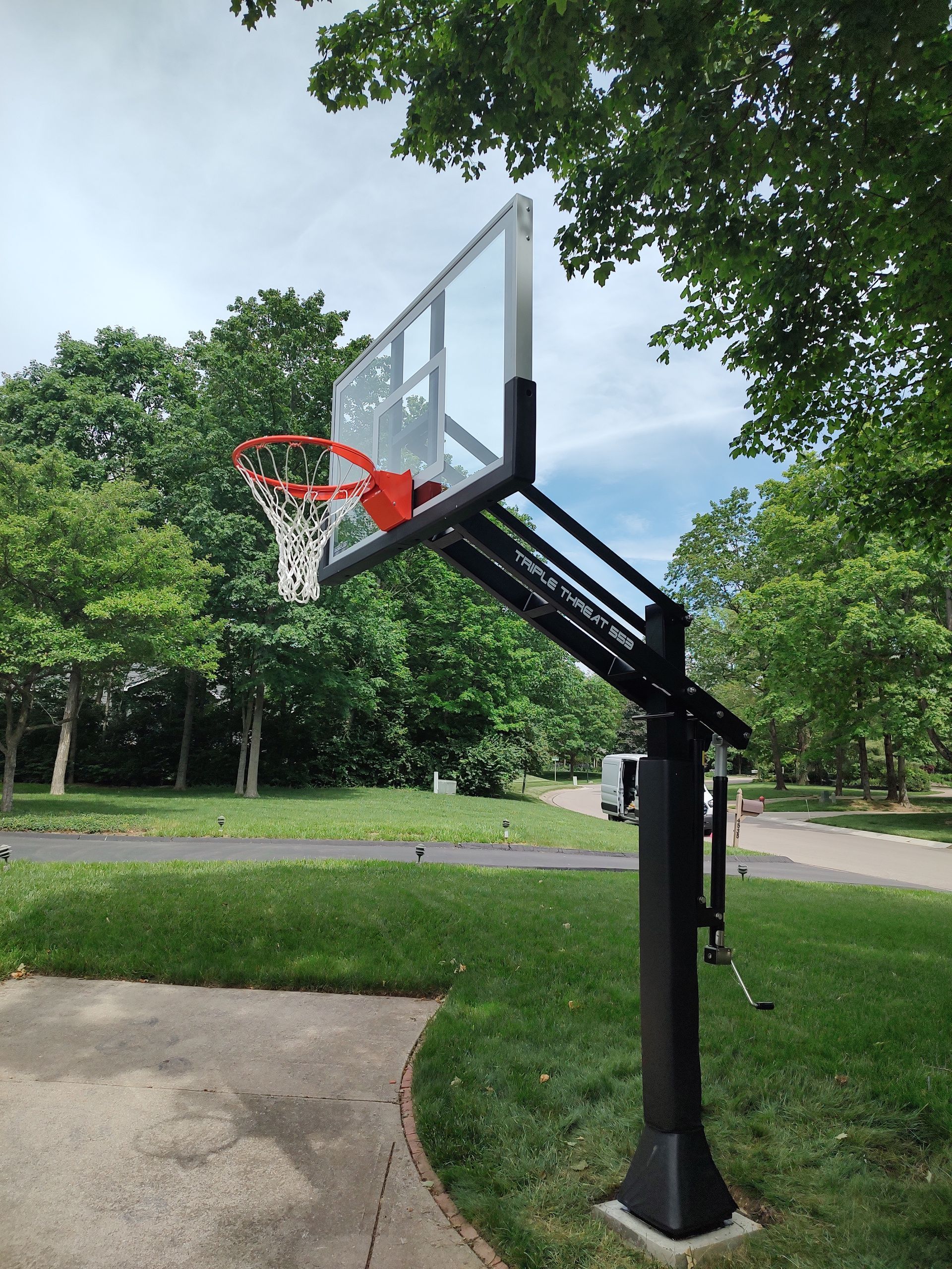 Portable outdoor basketball hoop beside a paved path in a park, with trees and grass around it