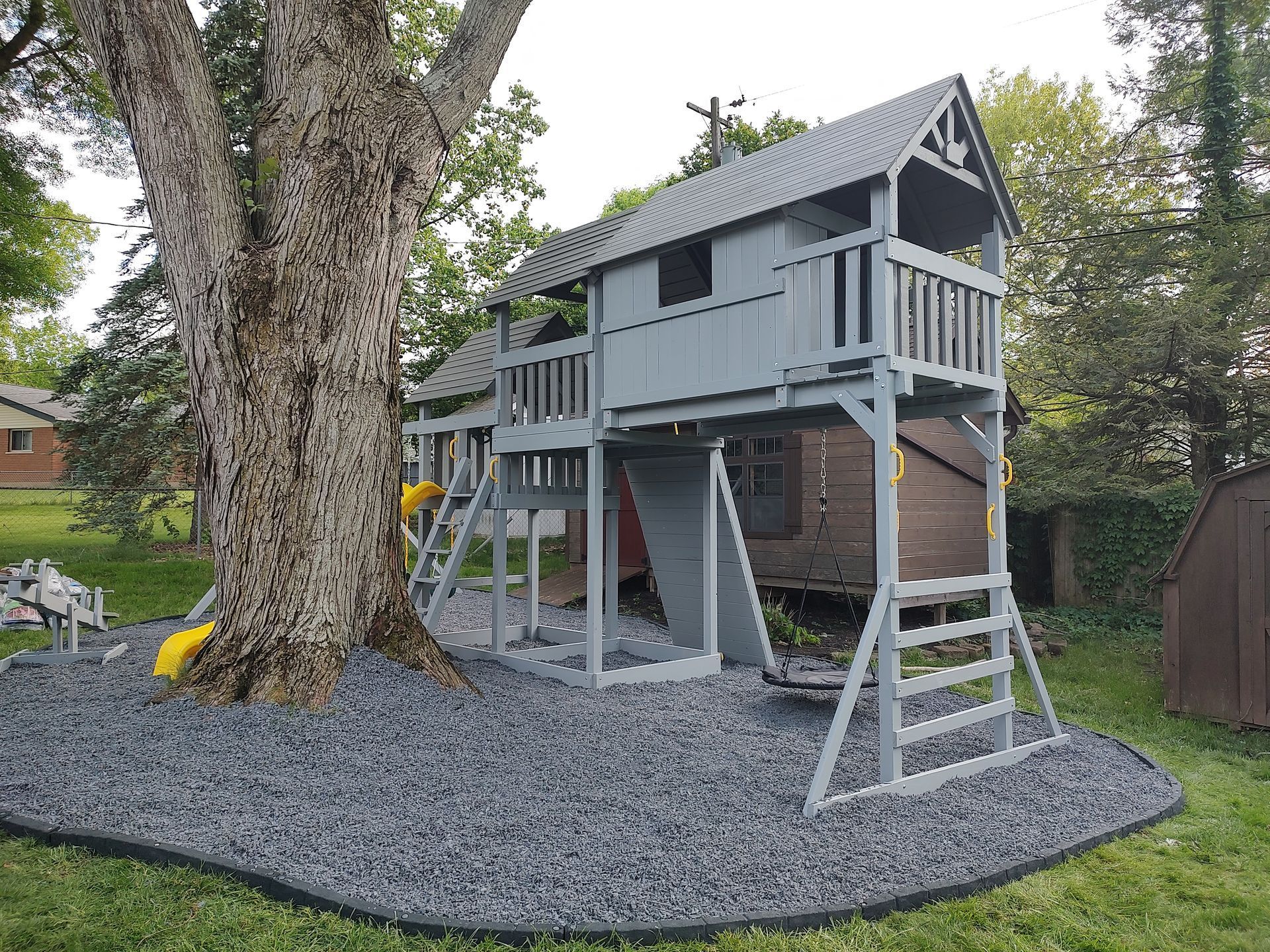 Treehouse playhouse on stilts in a backyard, beside a large tree and gray gravel ground cover.