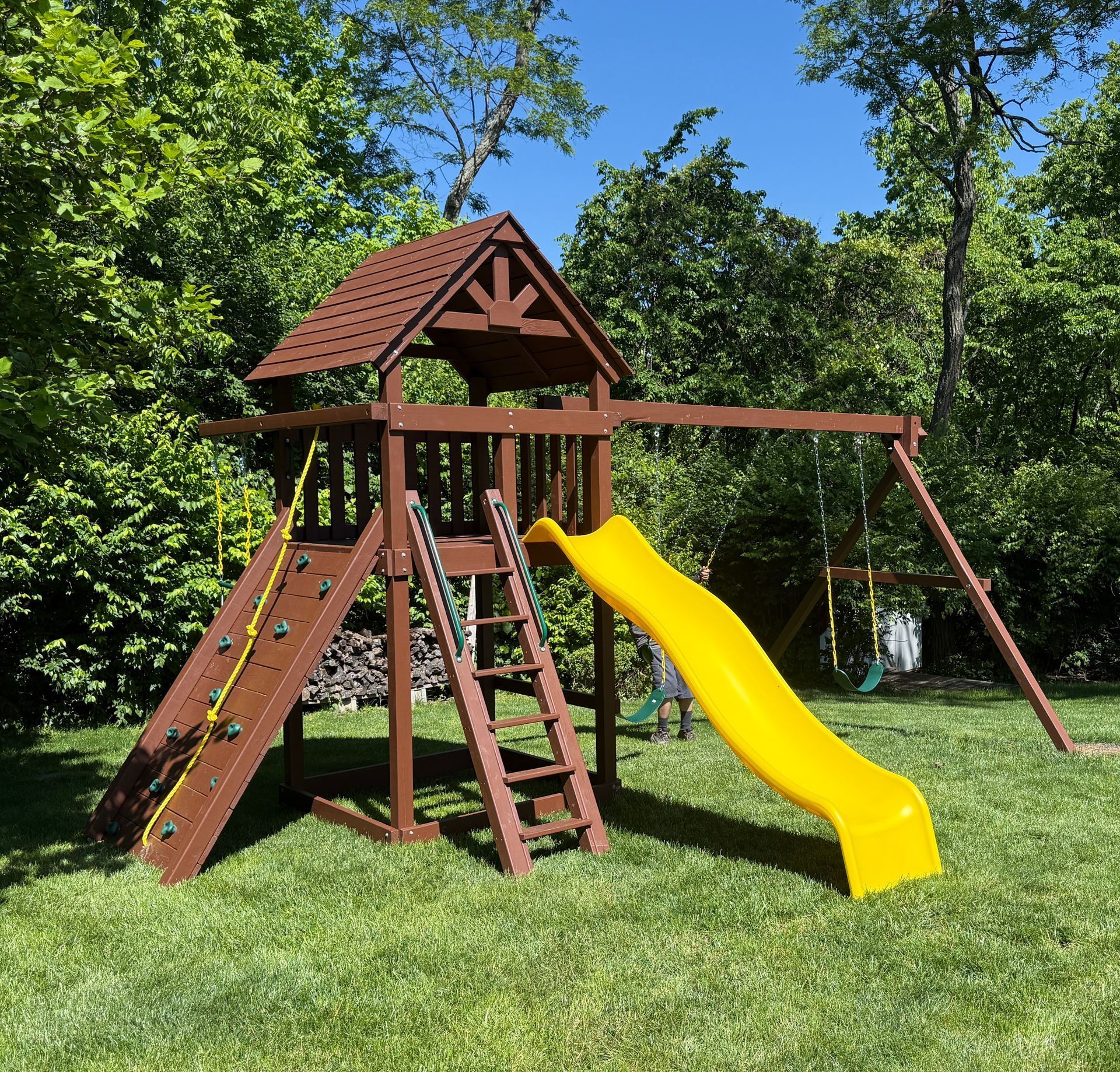 Wooden backyard playground with a yellow slide, climbing wall, and swing set on grass.
