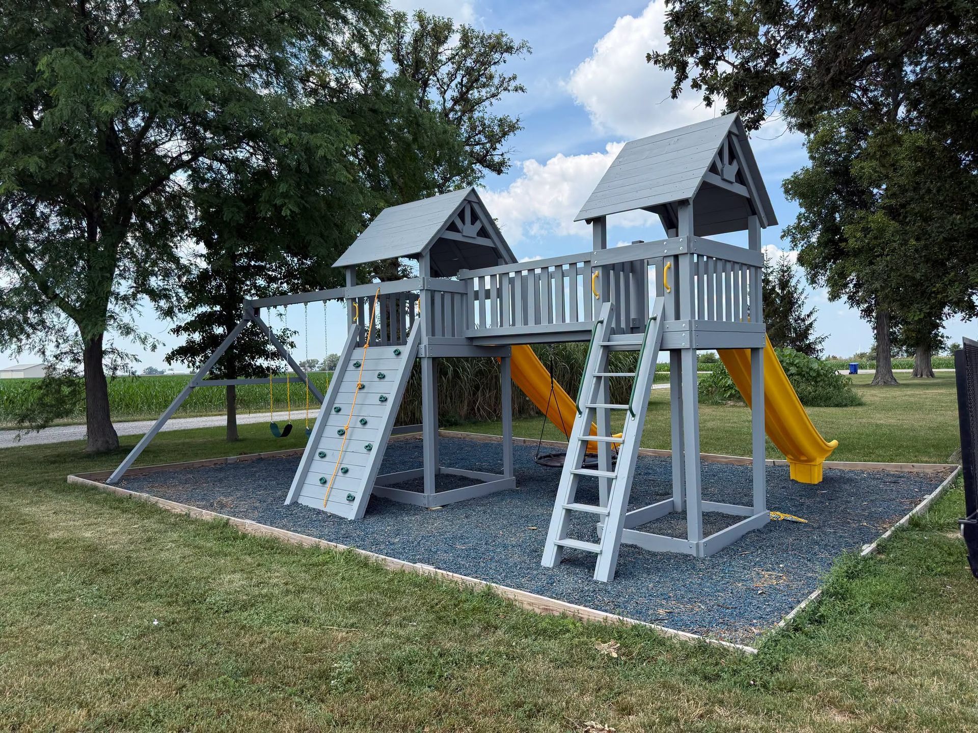 Children’s playground with a gray wooden climbing structure, slides, and swings on a grassy park lawn