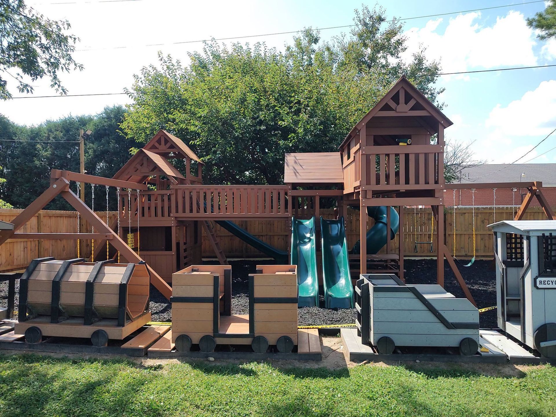 Wooden playground with slides, climbing structures, and play panels under a sunny sky.