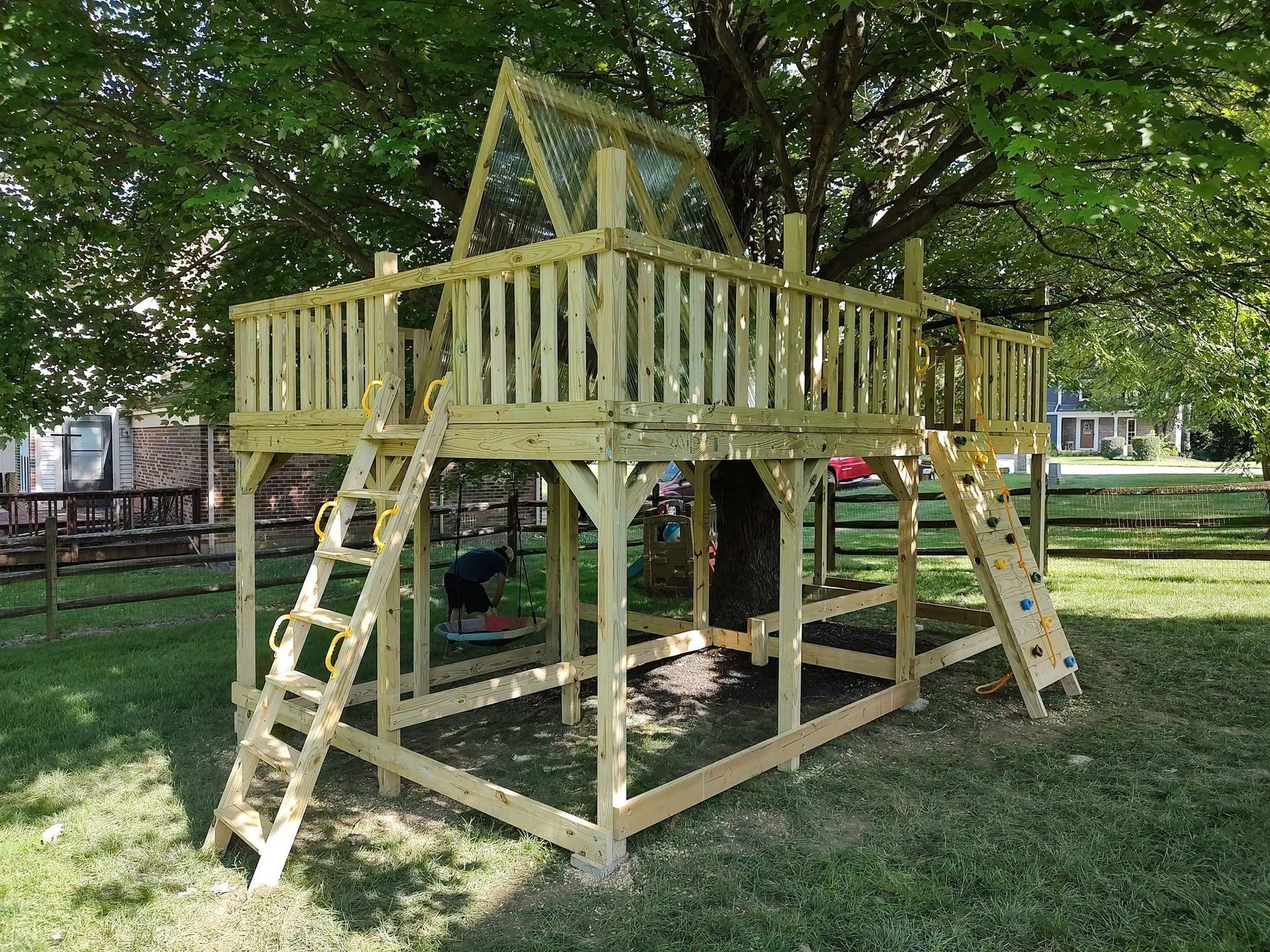 Wooden backyard play structure with deck, railings, and two ladders under trees.
