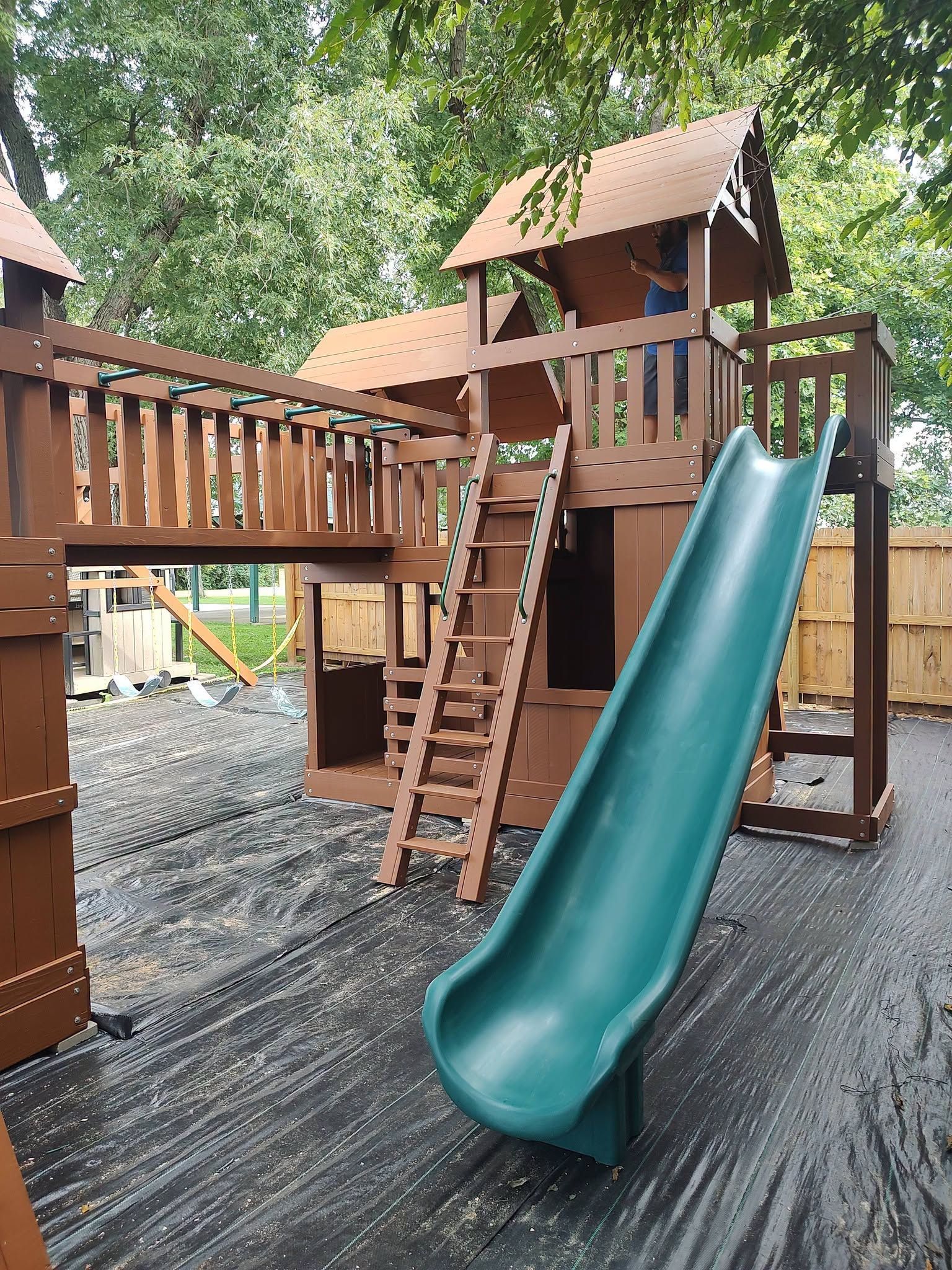 Wooden playground with a green slide and ladders on a rubberized surface under trees