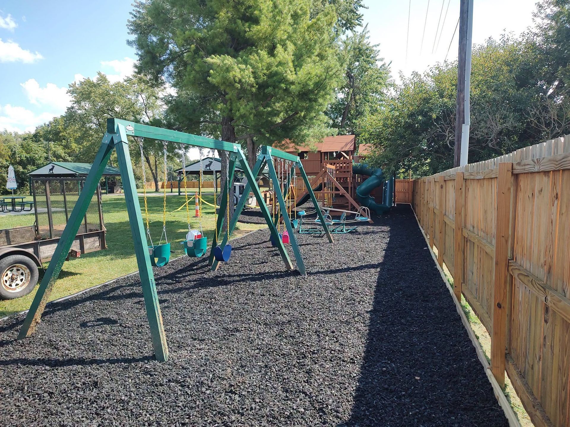 Playground with green swing set and wooden fence on a gravel surface, with trees and play structures in the background
