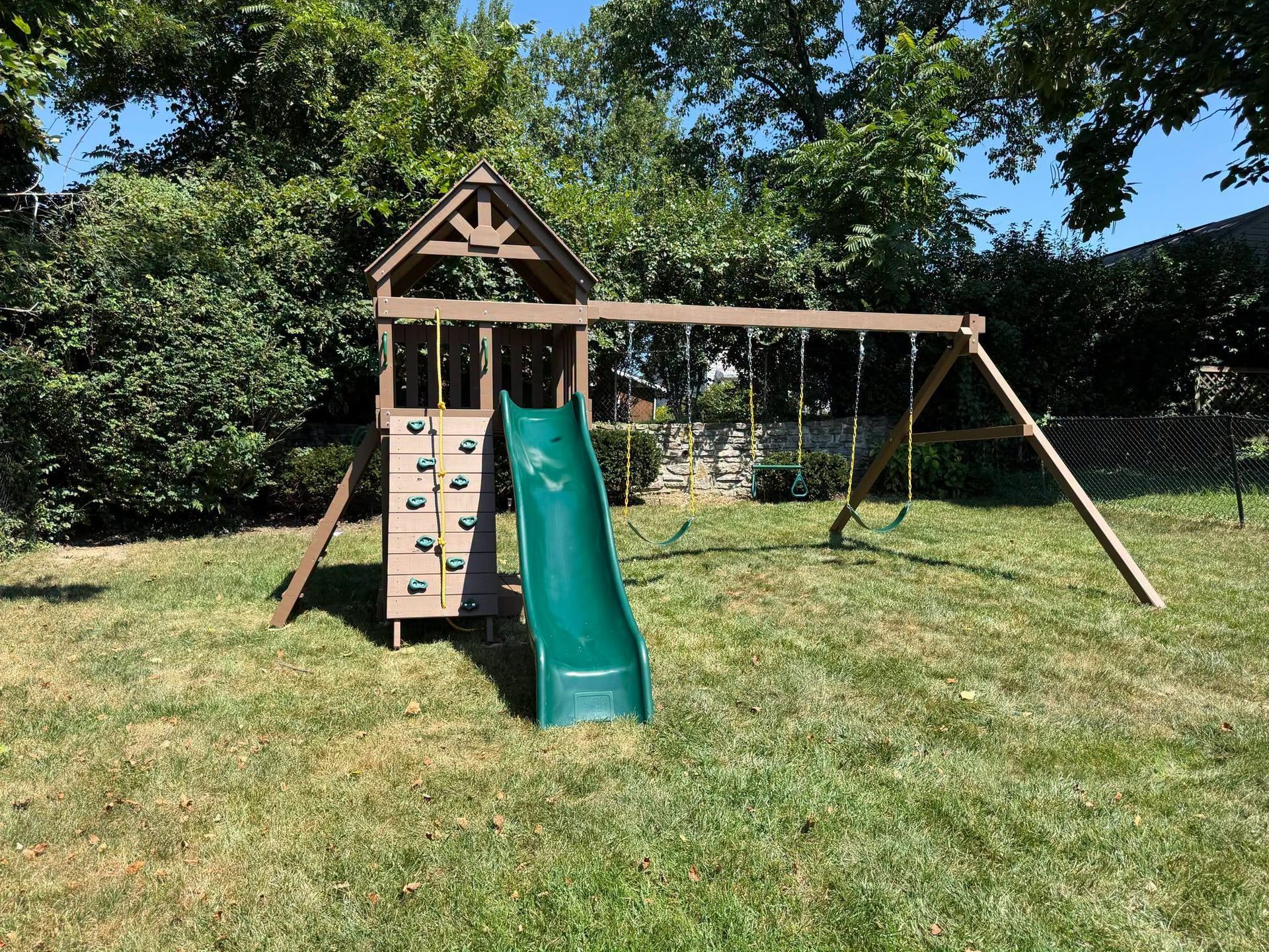 Wooden backyard playground with green slide, climbing wall, and swing set on a grassy lawn.