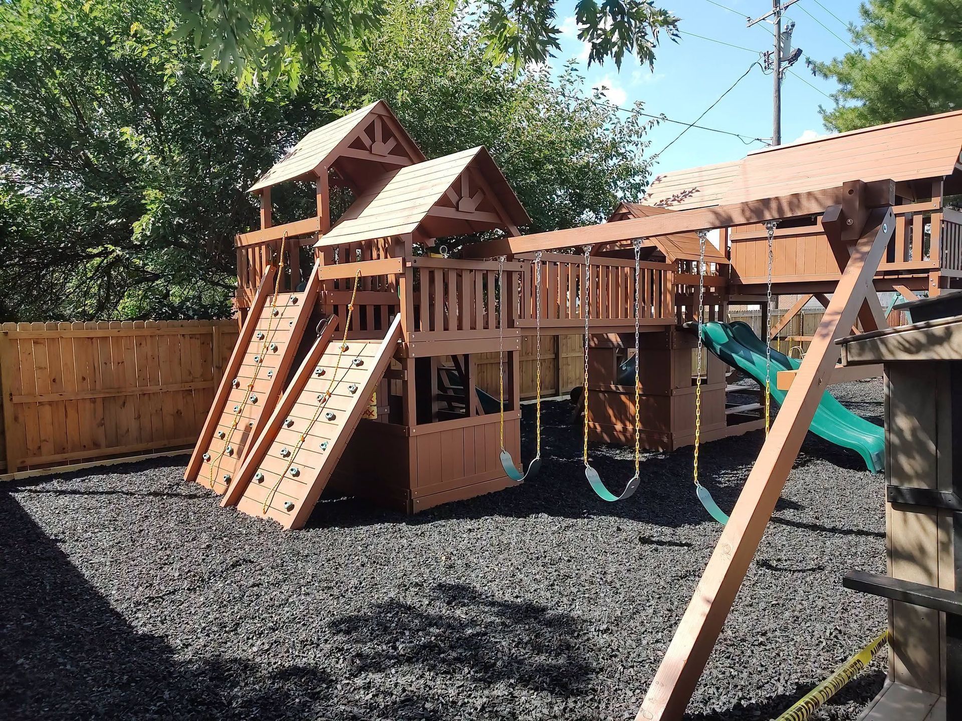 Wooden backyard playset with slides, climbing wall, and swing set on black gravel mulch.
