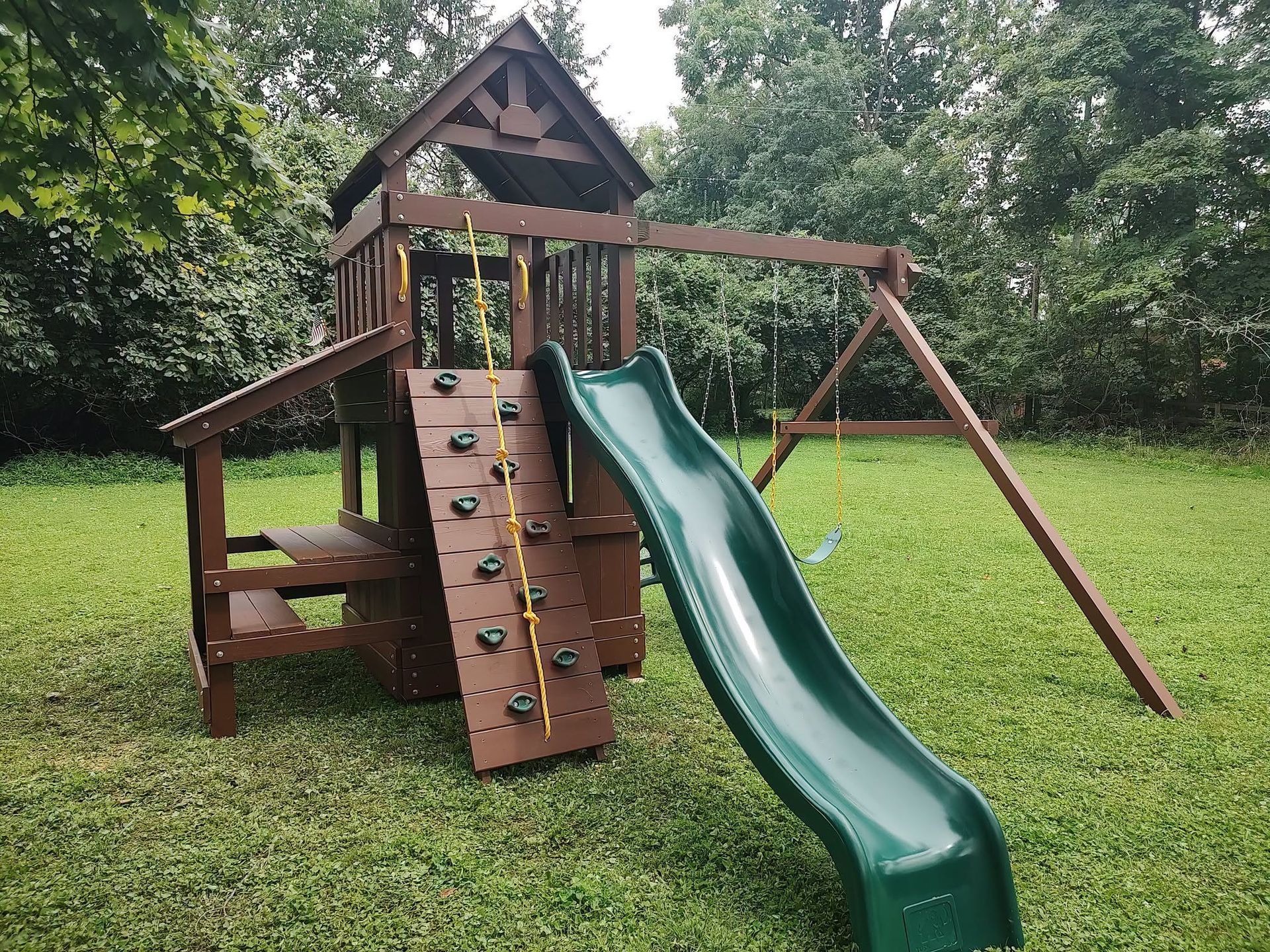 Wooden backyard playset with green slide, climbing wall, and swing set on grass.