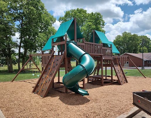 Playground structure with teal slides and wooden stairs in a grassy park under a sunny sky