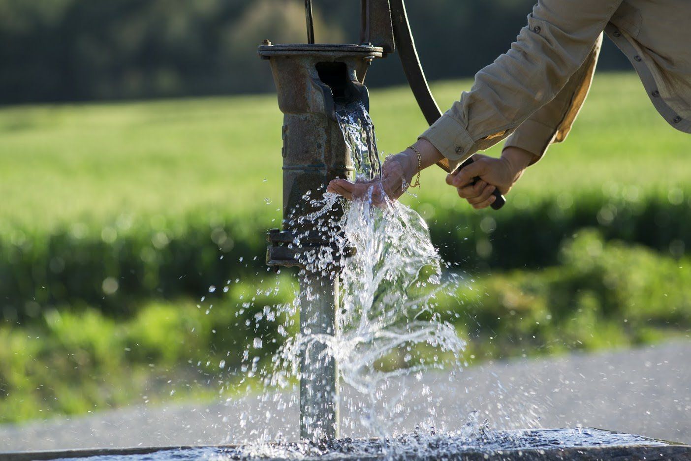 Water flows from a rustic hand pump over someone’s hands against a blurred green field.
