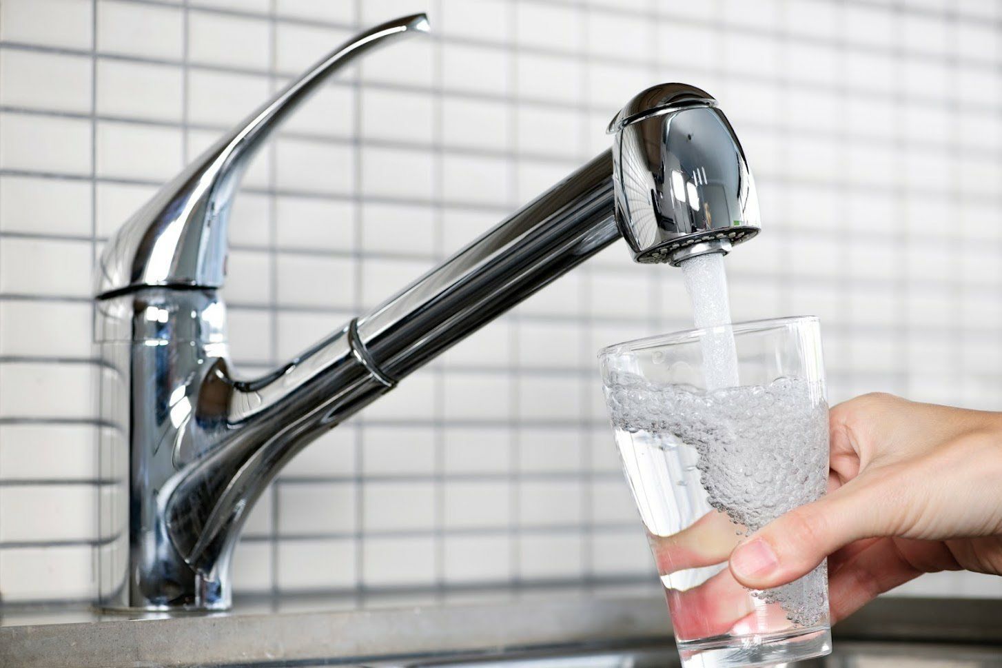 A hand holds a glass under a chrome kitchen faucet as water fills the glass in front of a white tiled backsplash.