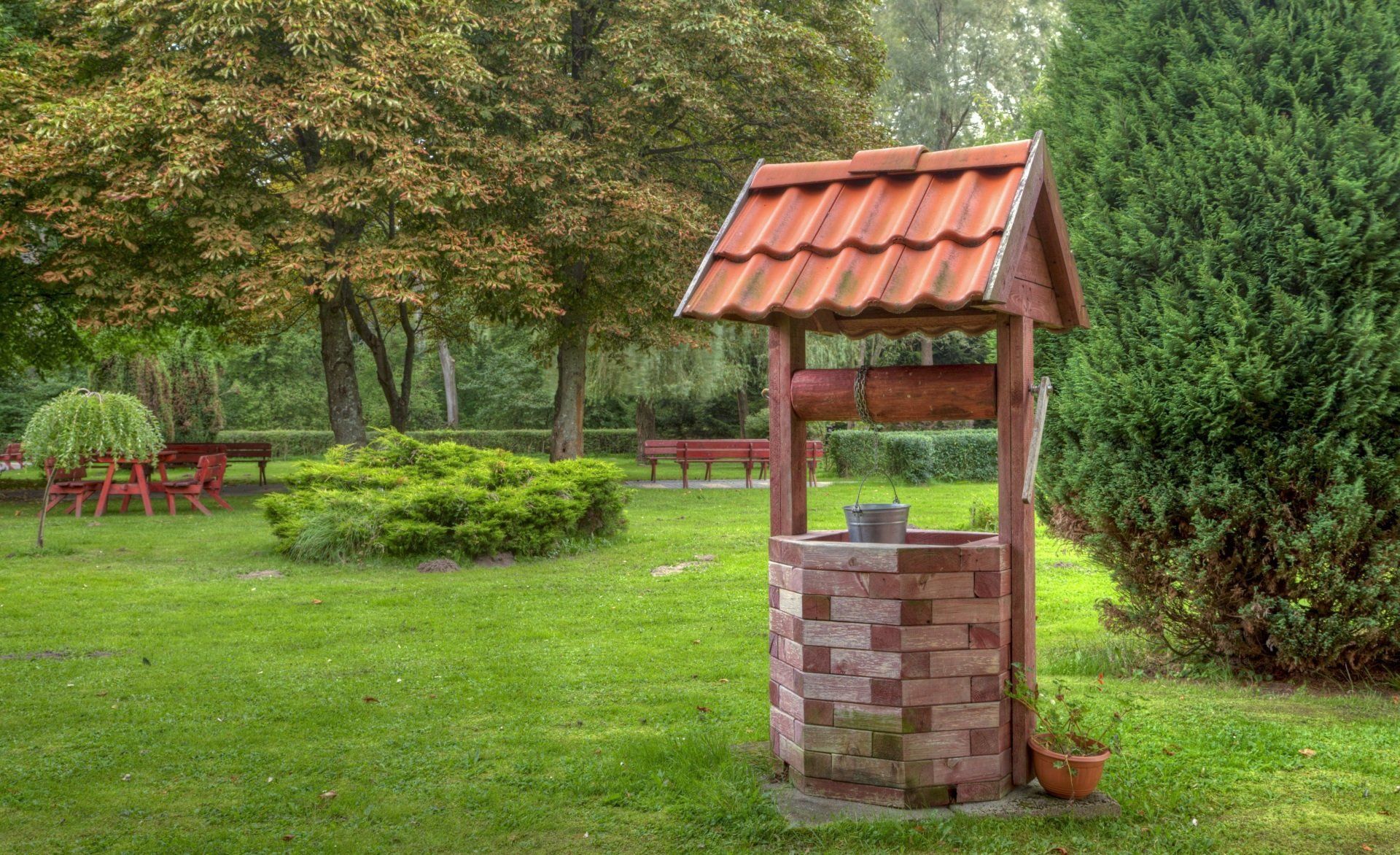 A decorative brick garden well with a red-tiled roof stands on a grassy lawn surrounded by trees and shrubs.