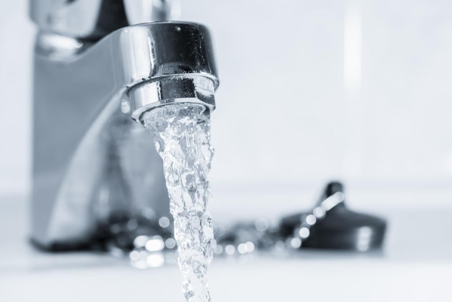 A chrome faucet with a stream of water flowing from it into a sink, with a sink stopper and chain visible in the background.