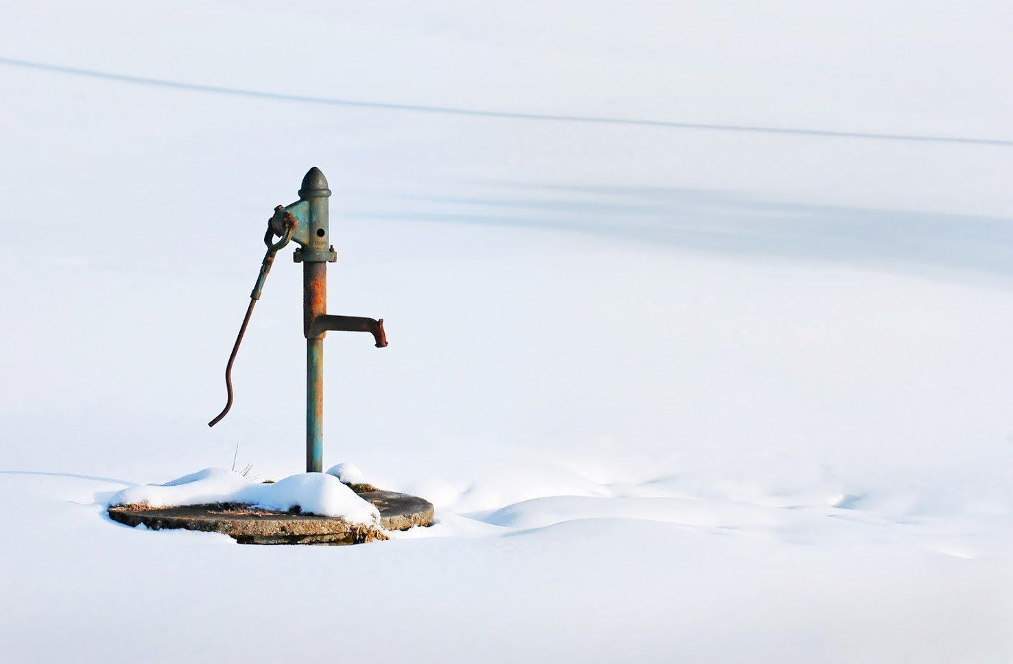 A weathered, vintage hand water pump stands alone in a vast, flat, snow-covered landscape.