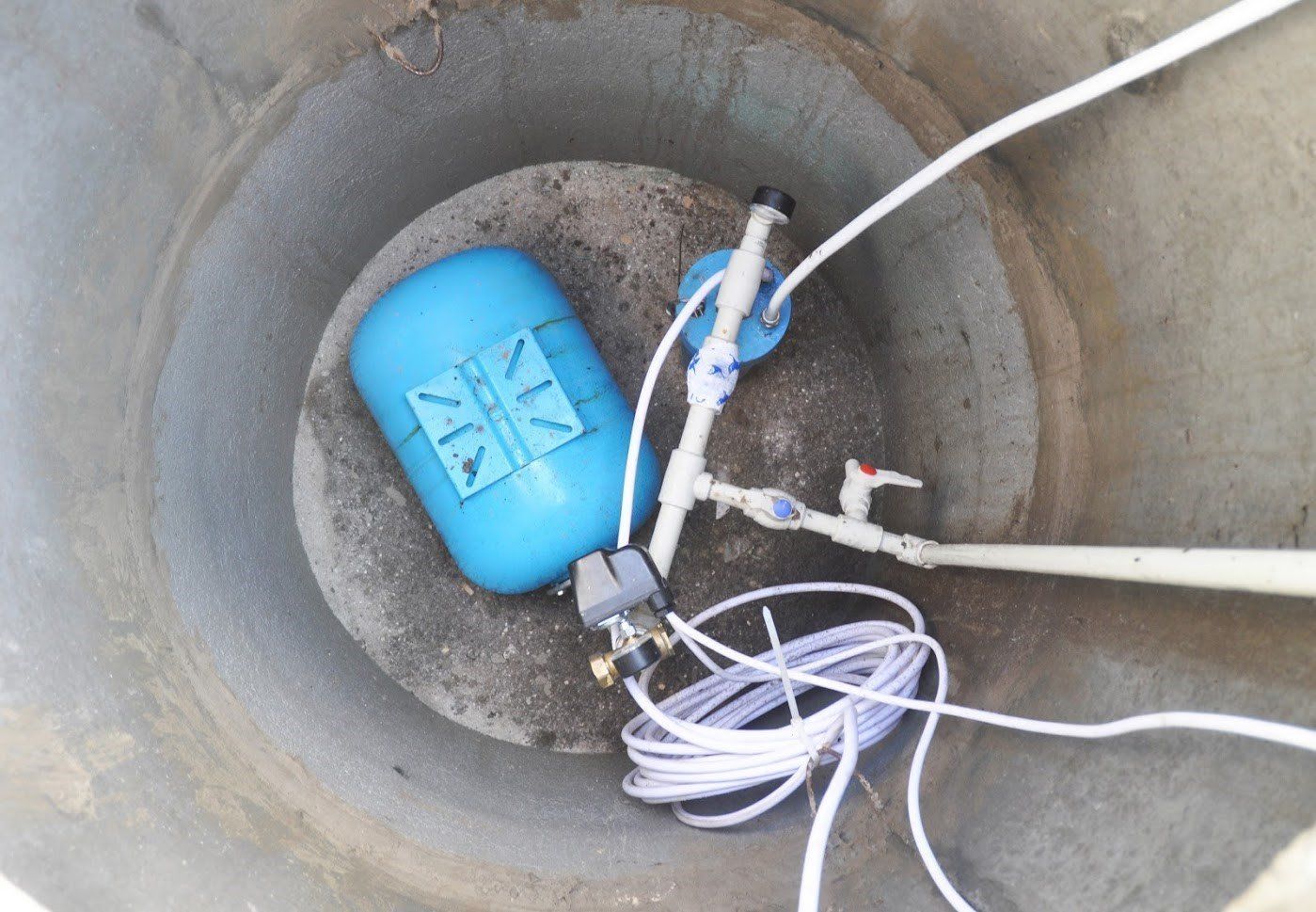 A blue pressure tank and pump assembly with white piping installed inside a cylindrical concrete well casing.