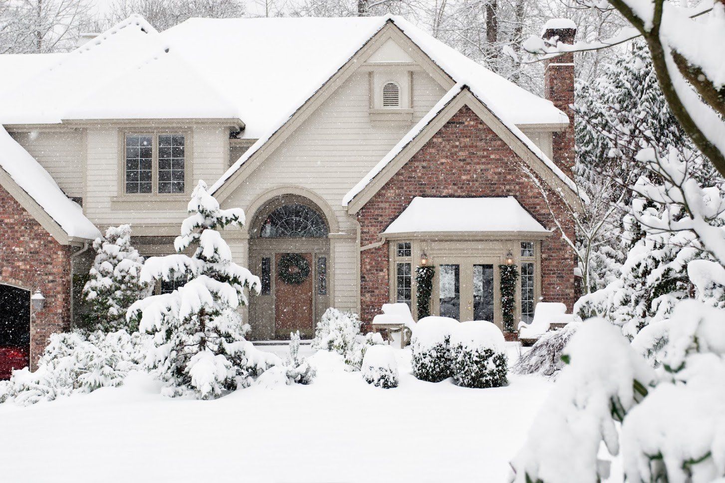 A two-story brick and siding house covered in a thick blanket of winter snow with snow-covered trees and bushes in front.