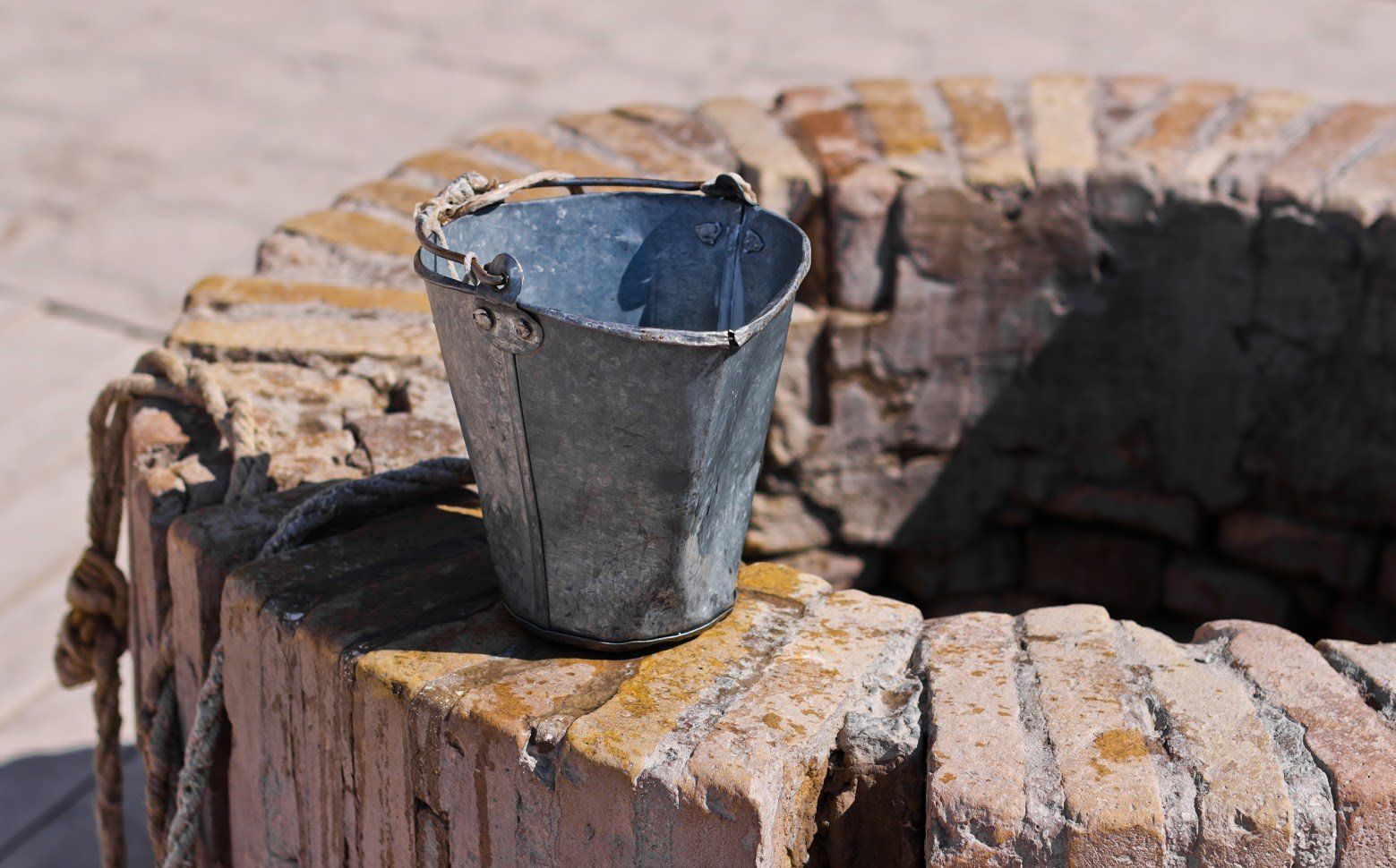 A weathered metal bucket sits on the edge of a rustic, circular brick well.