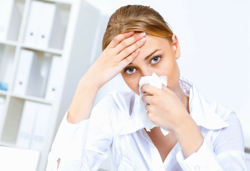 A person in a white shirt holding a tissue to their nose with one hand and resting the other on their forehead.