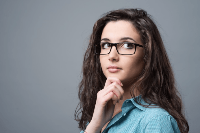 A person with wavy dark hair wearing glasses and a blue shirt, looking away with a hand resting thoughtfully on their chin.