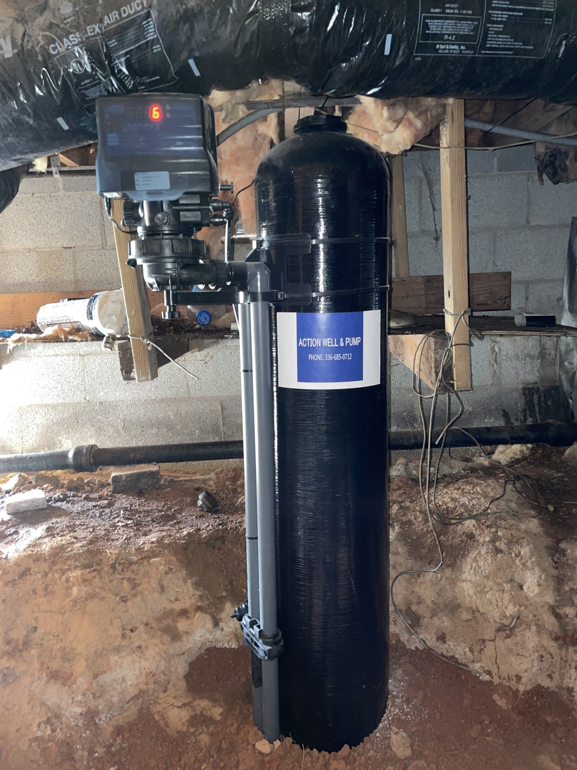 A water filtration tank with a digital control head installed in a crawl space with exposed insulation and dirt flooring.