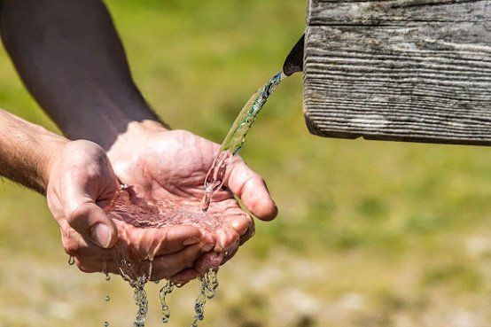Cupped hands catching fresh water flowing from a rustic wooden spout in a natural, outdoor setting.