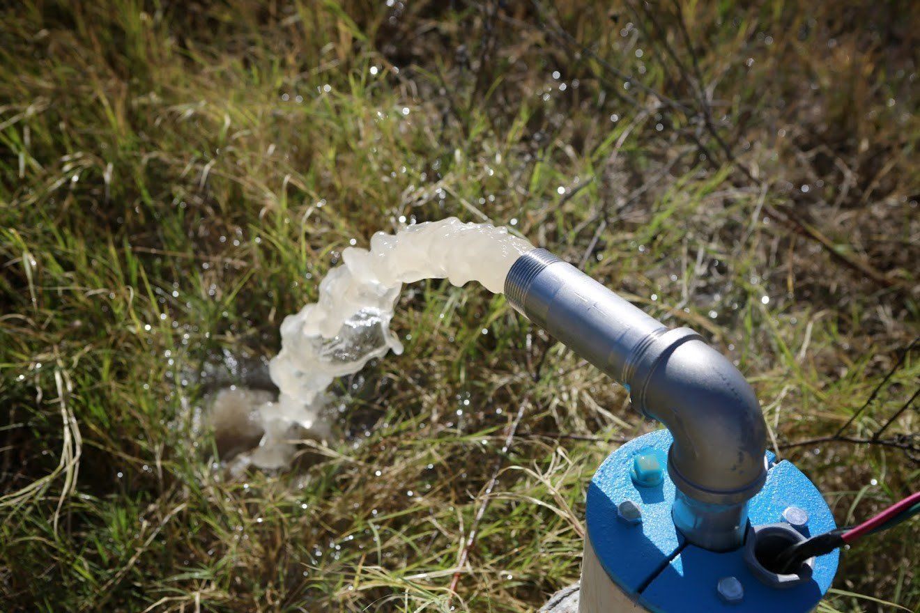 Water flows from a metal pipe mounted on a blue cap into grassy ground, likely from a well pump.