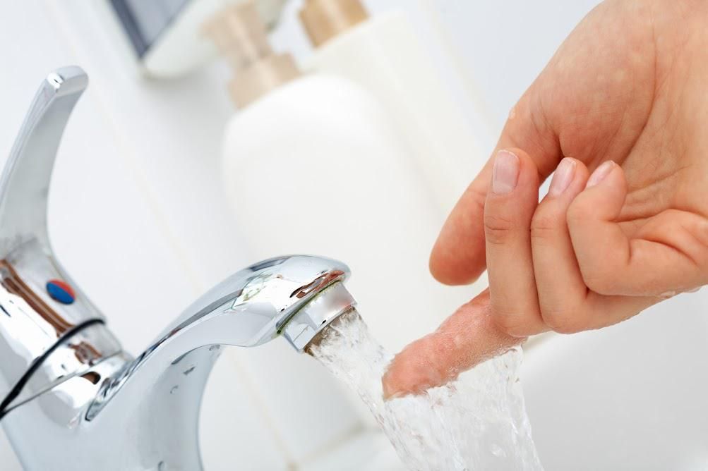 A hand tests the temperature of water flowing from a silver bathroom faucet next to soap dispensers.