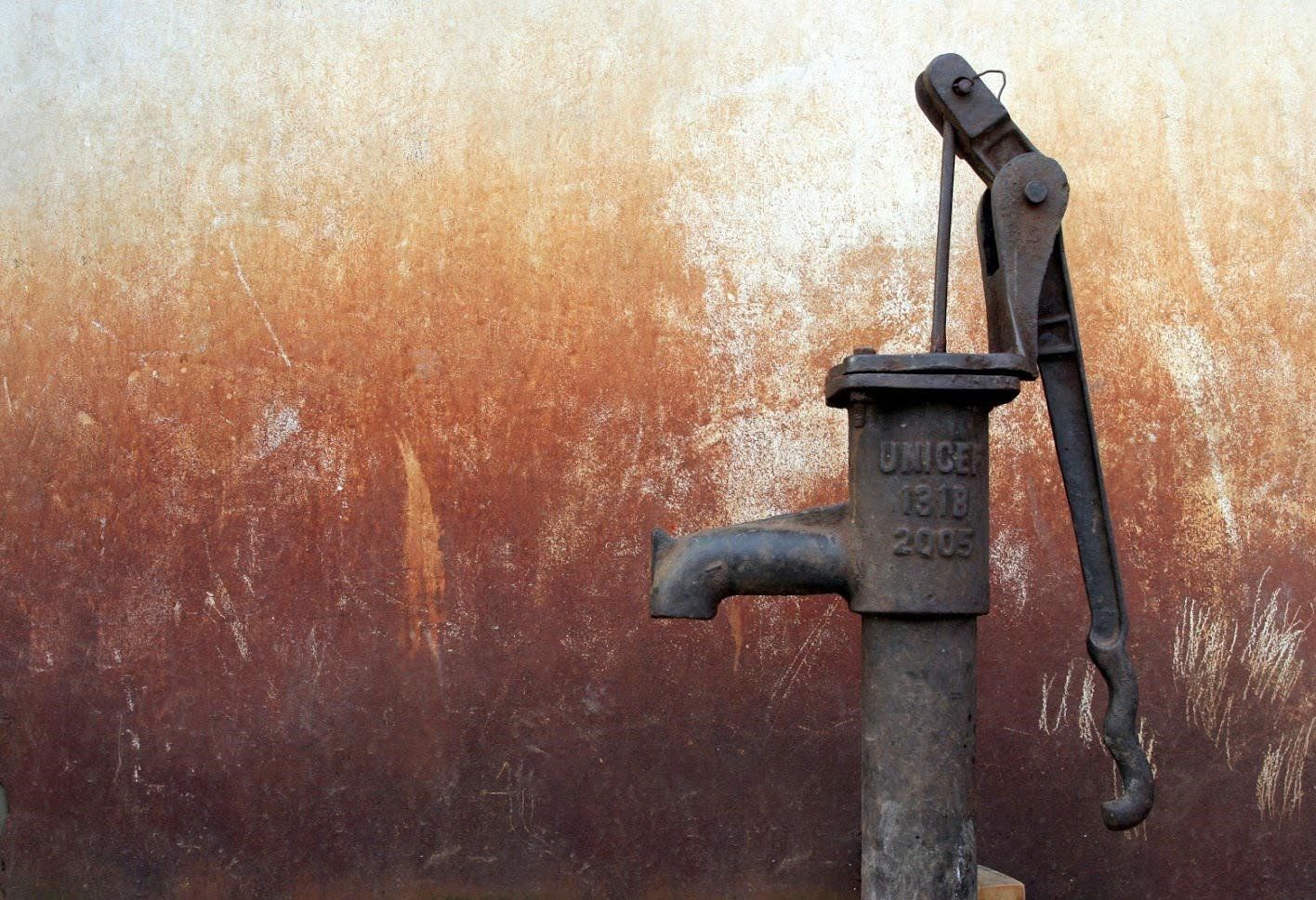 A weathered, dark metal hand pump stands against a textured, rust-colored wall.