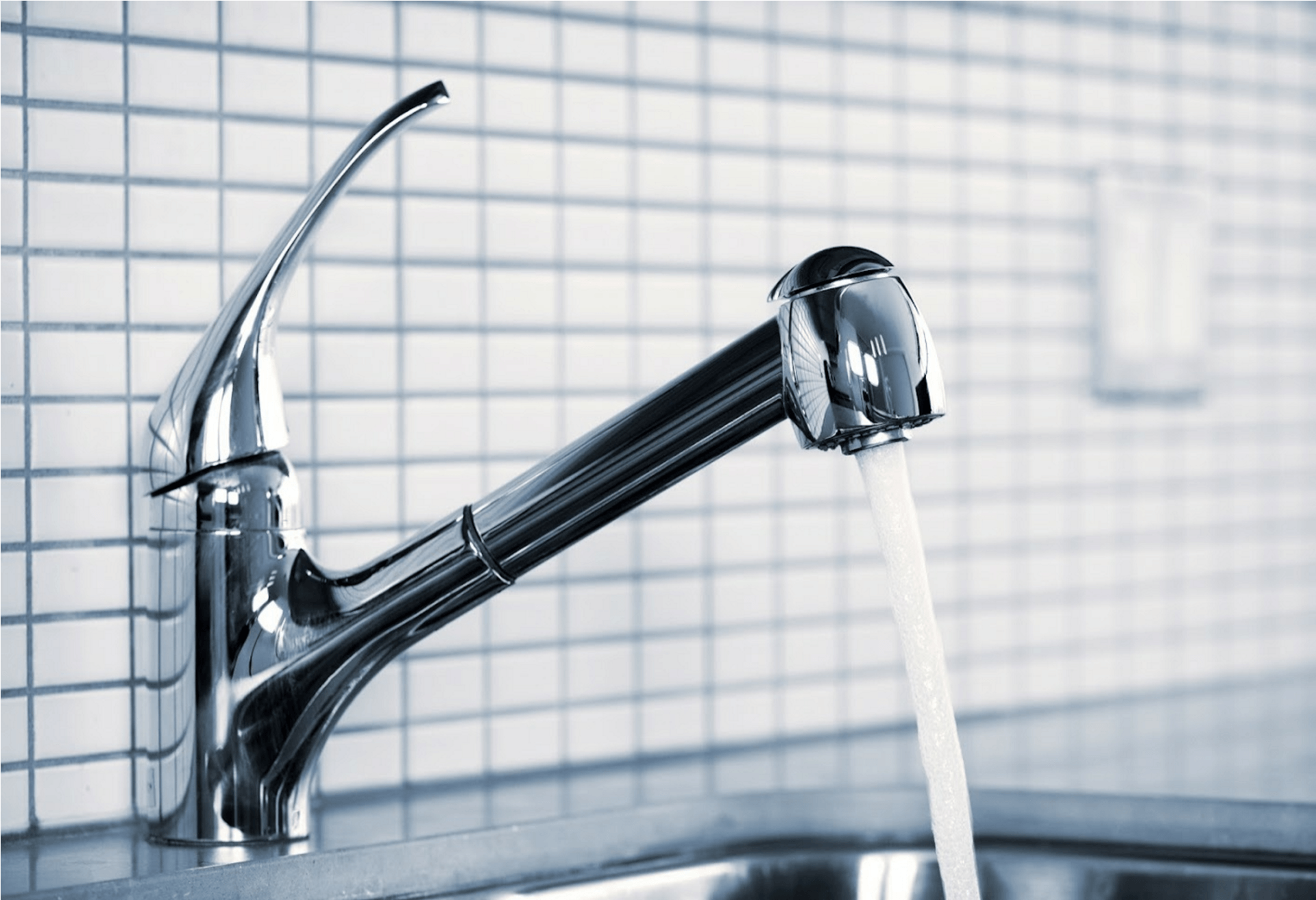 A chrome pull-down kitchen faucet with water running, set against a white tiled backsplash.