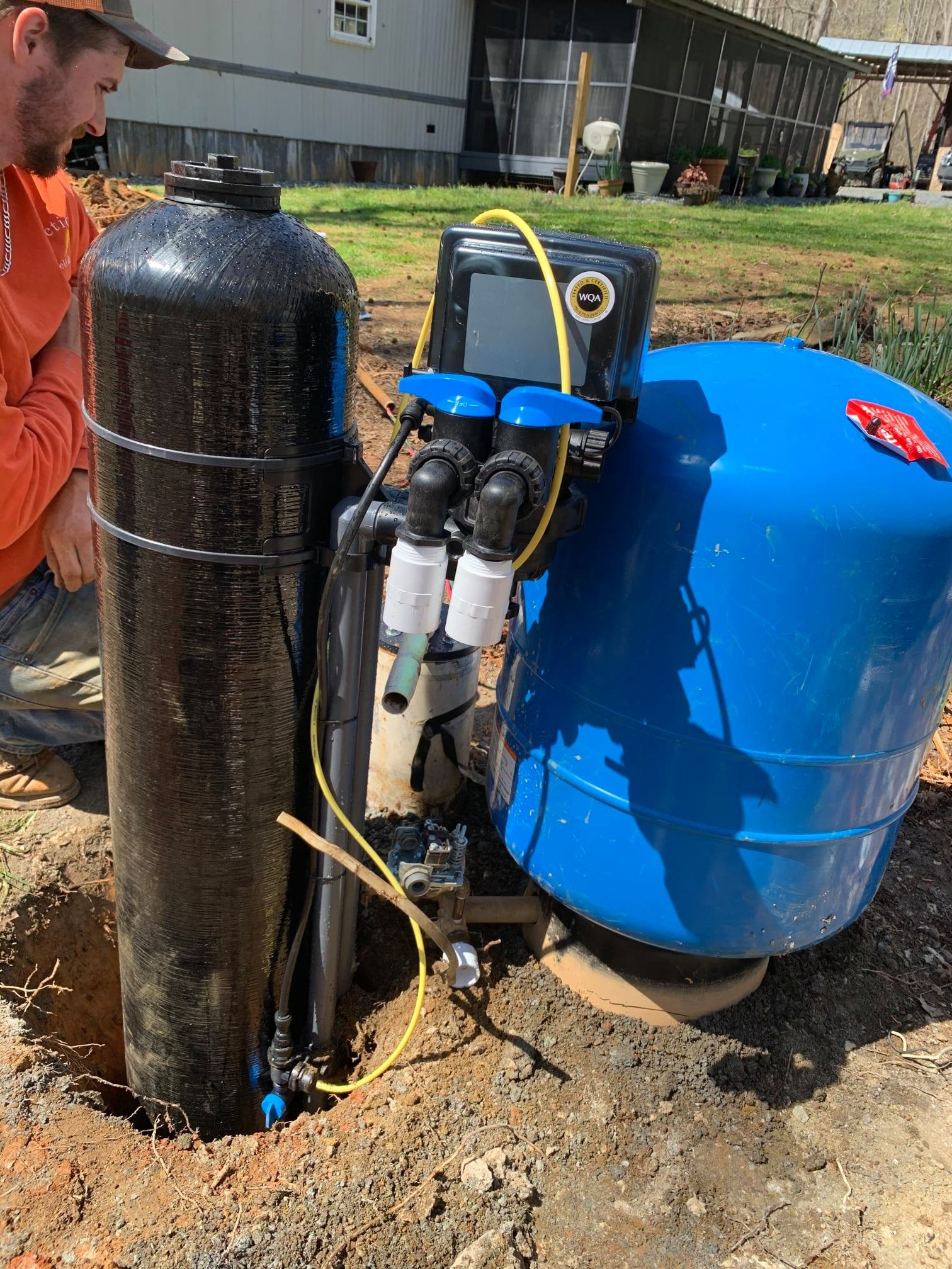 A worker installs a black water filtration tank and a large blue pressure tank outdoors on a dirt ground near a home.