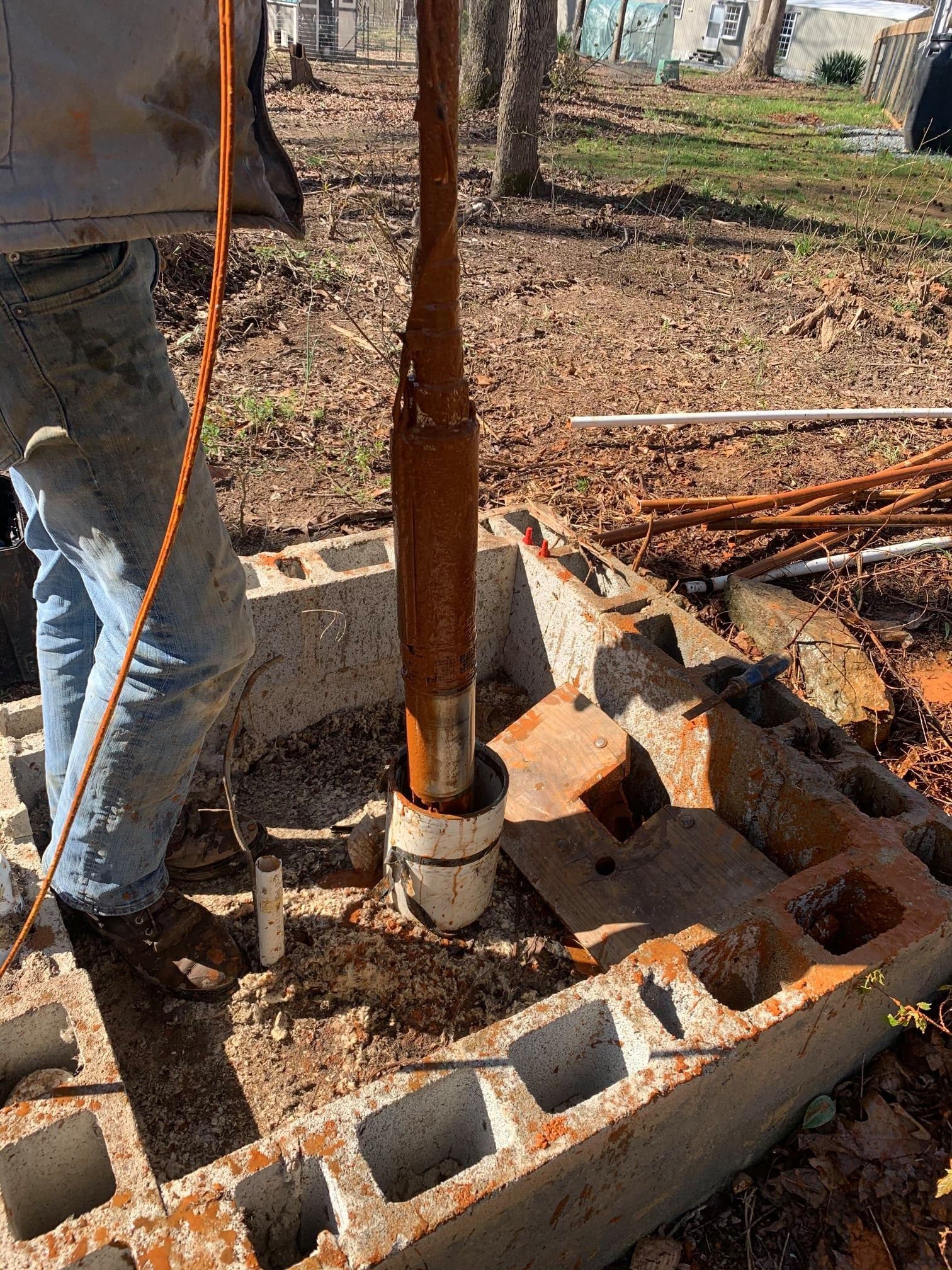 A rusted well pump being pulled out of a PVC casing inside a concrete block structure in a yard.