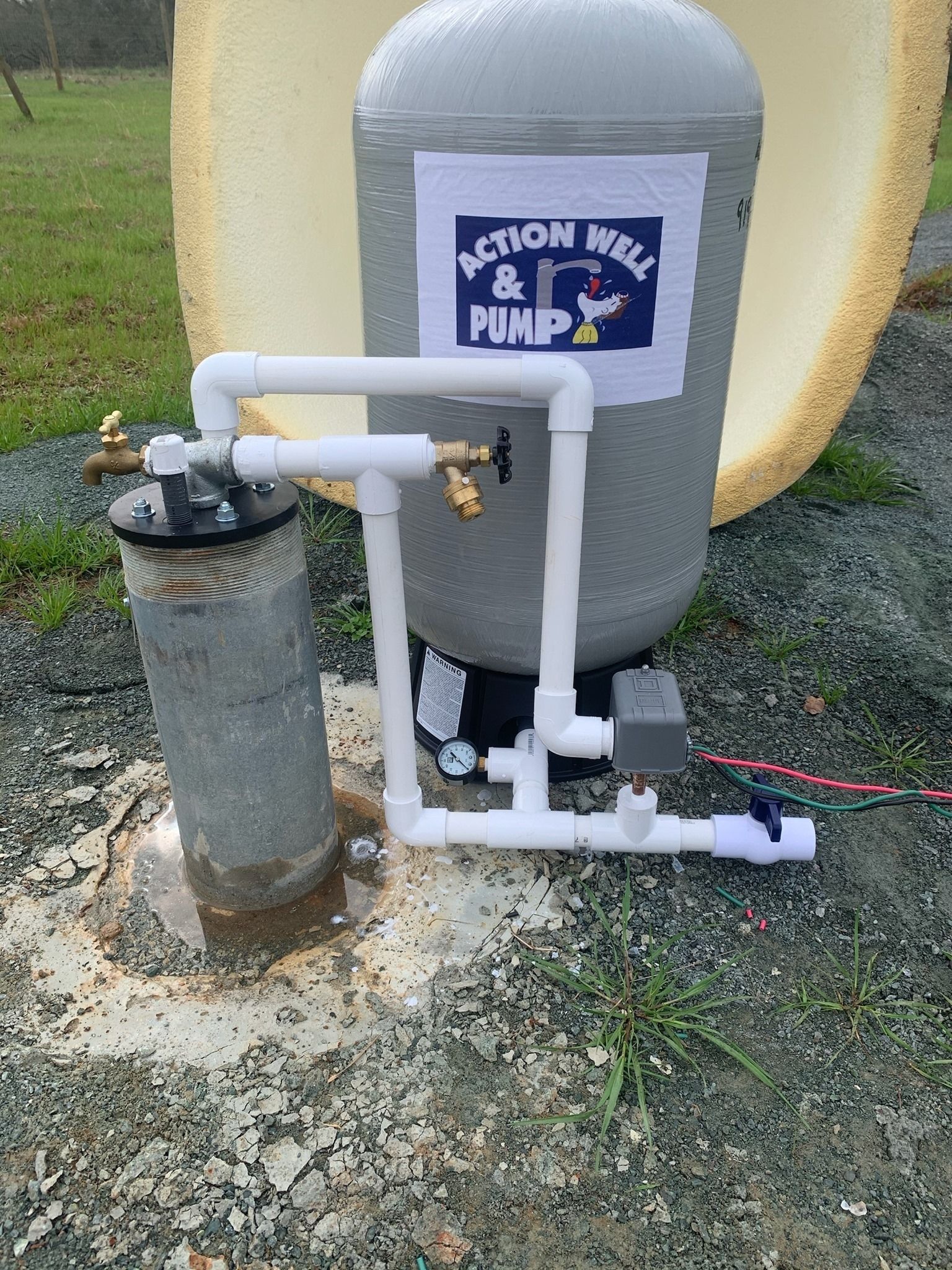 A water well pump installation featuring a gray pressure tank with an Action Well & Pump logo next to a PVC-piped wellhead.
