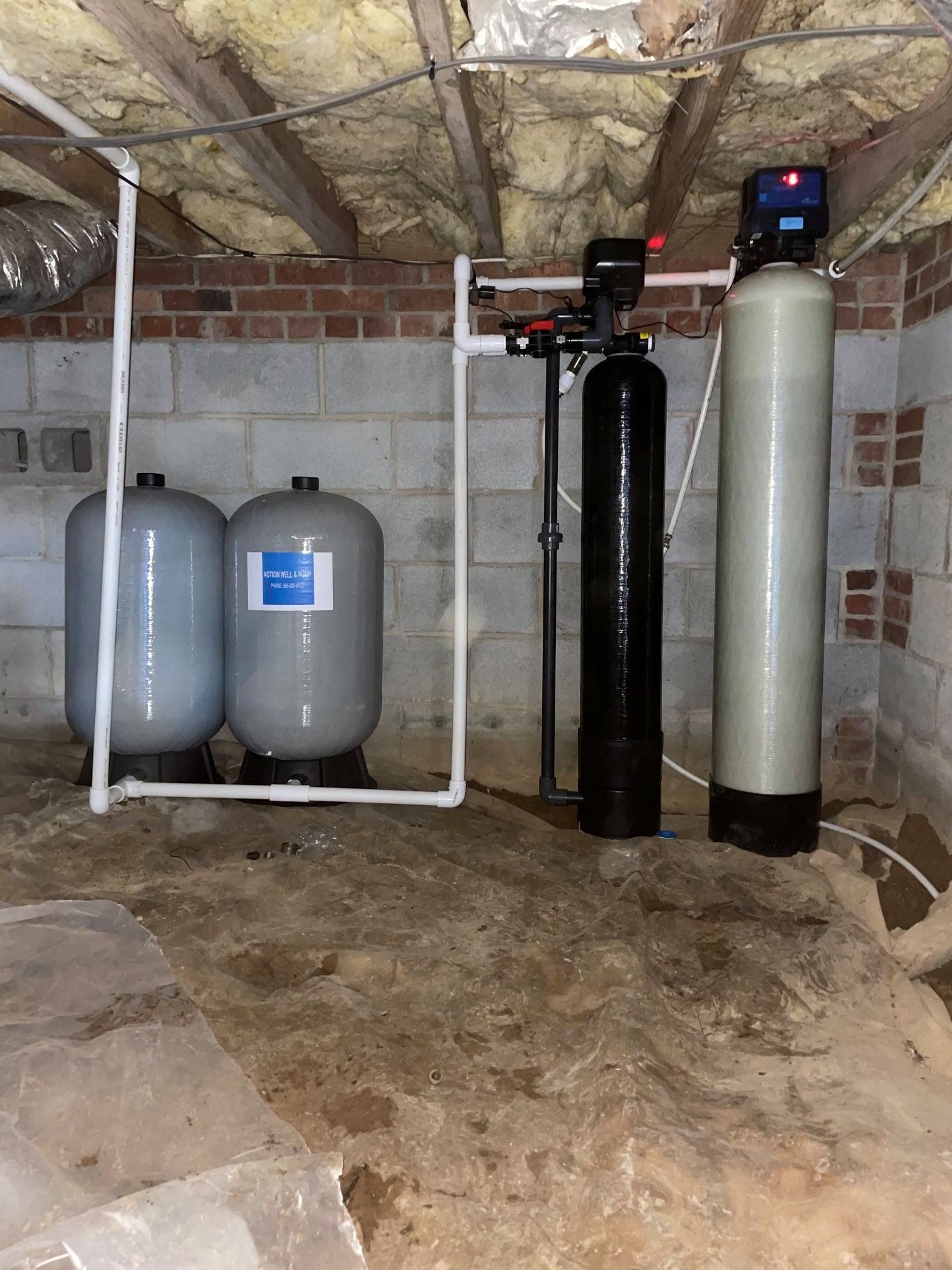 Water treatment system with two grey tanks, a black filter, and a tall white tank in a basement with brick walls.