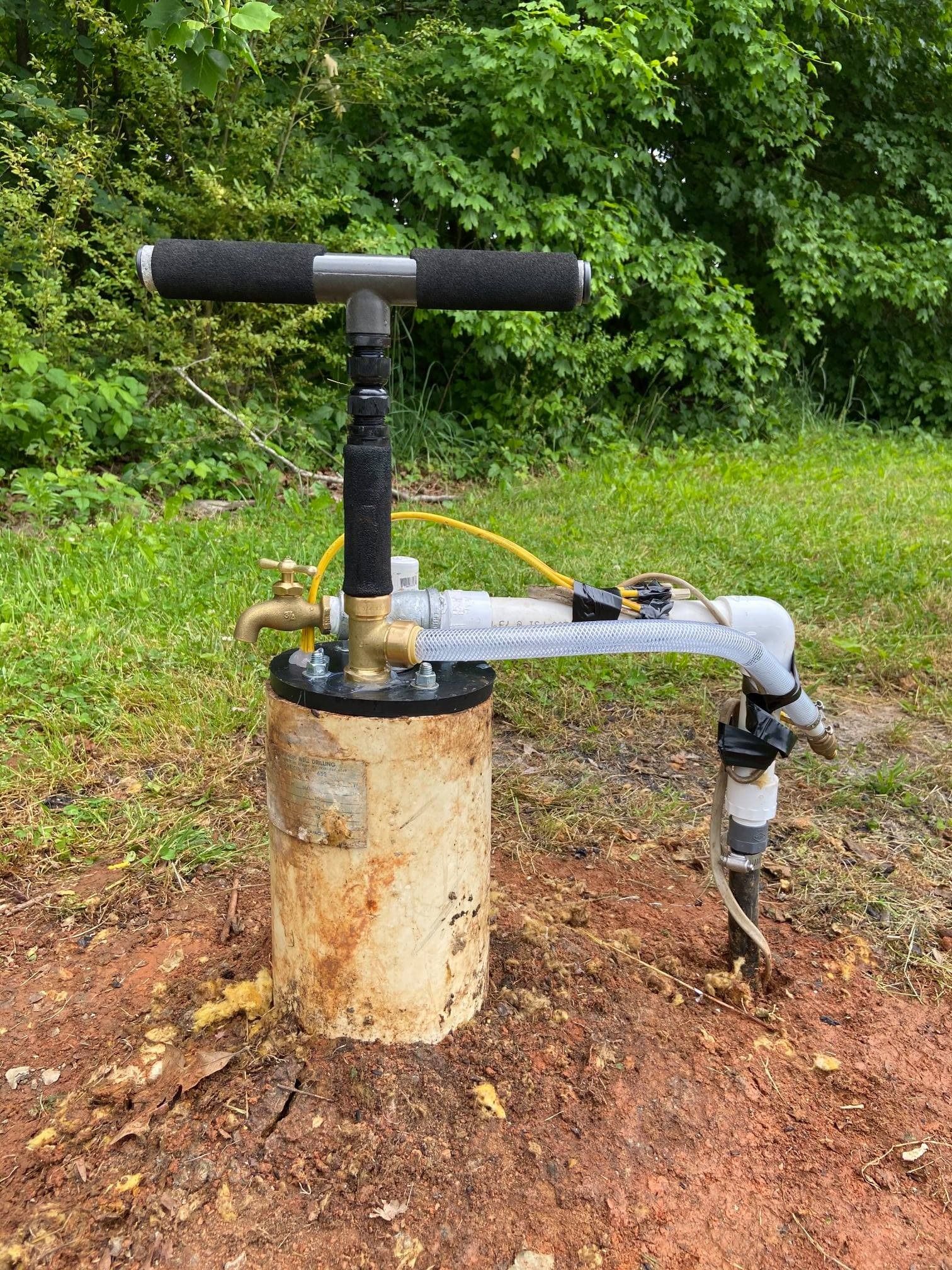 A hand-operated water pump mounted on a well casing in a grassy, wooded area.