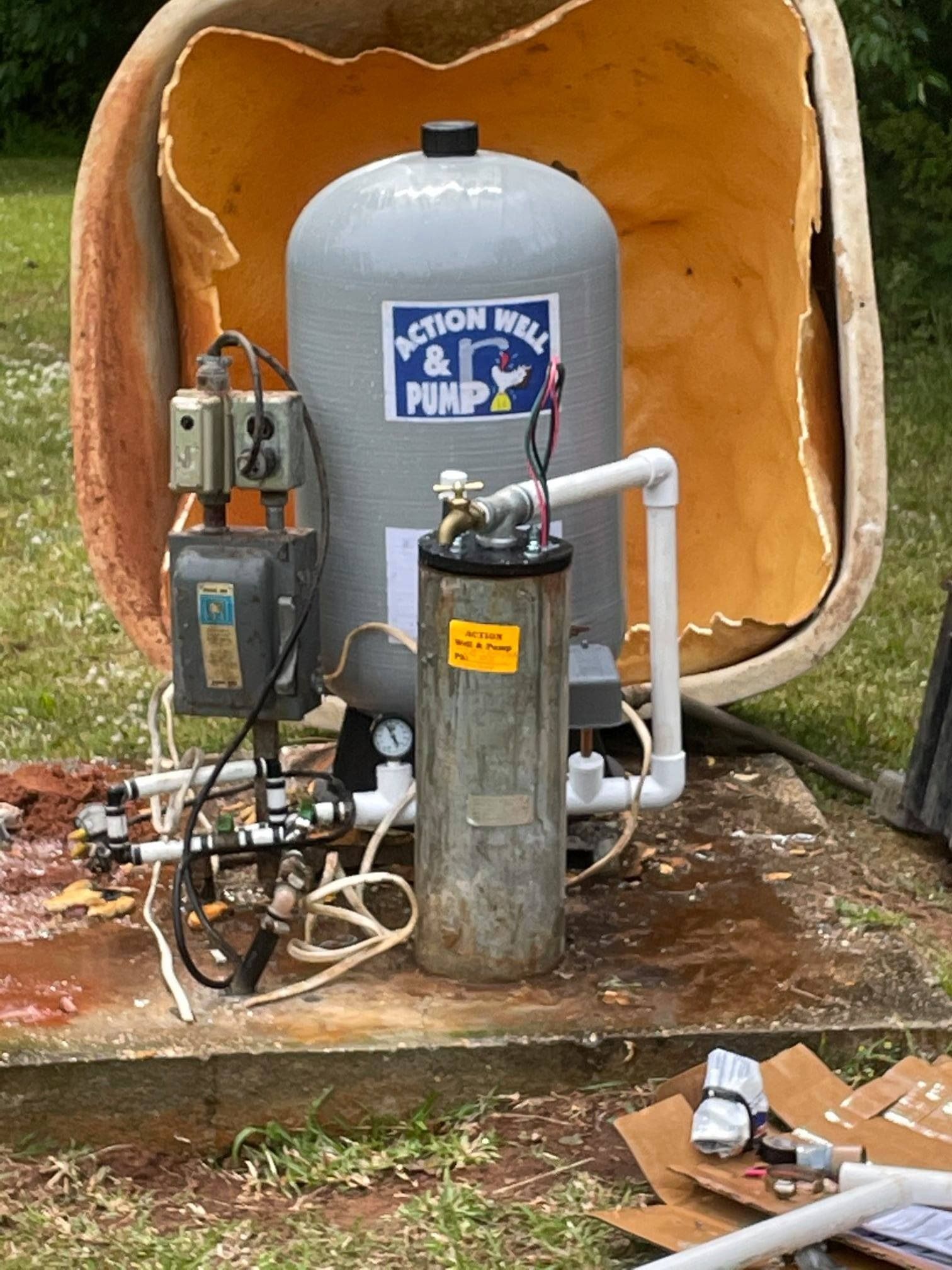 A grey well pressure tank and a metal water filtration unit sit on a concrete pad outdoors, with a protective cover behind.