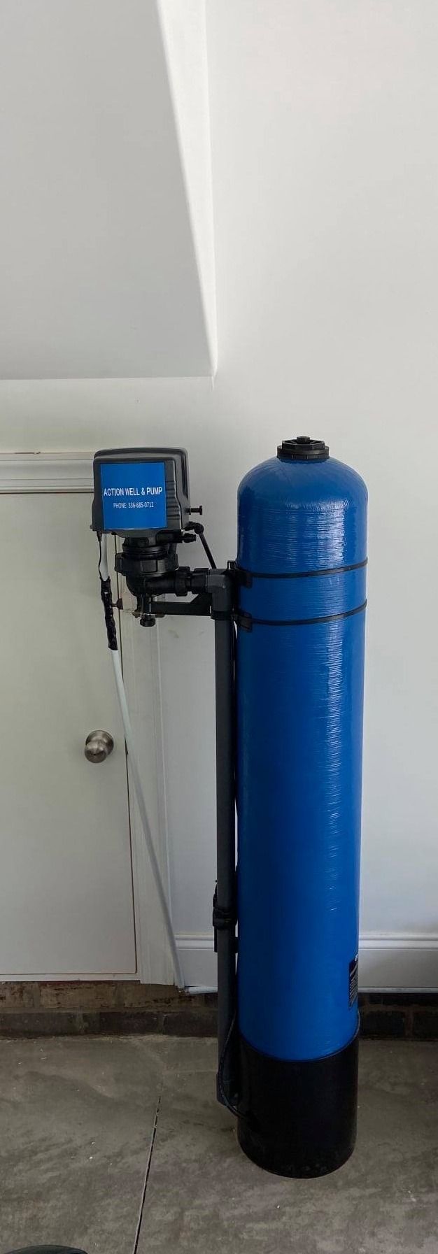 A tall, blue water softener system with a digital control valve stands against a white wall next to a white door.