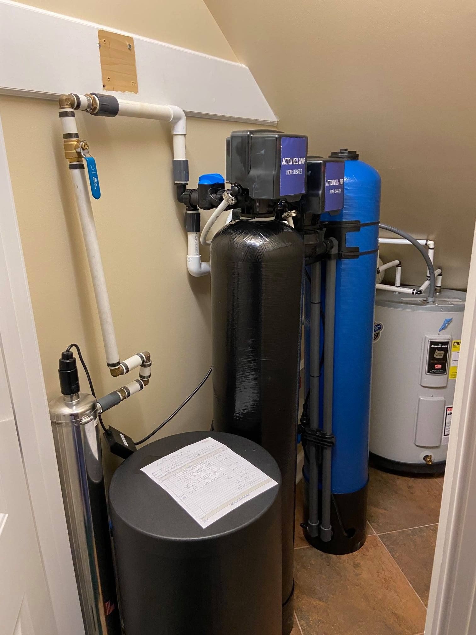 Water filtration and softening equipment in a utility closet, including black and blue tanks, pipes, and a water heater.