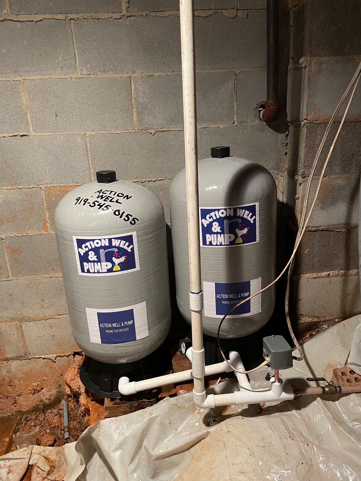 Two gray well water pressure tanks connected by white PVC pipes against a concrete block wall in a basement area.