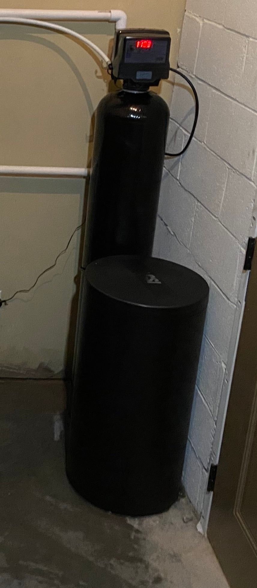Black water softener system with a digital control head installed against a concrete block wall.