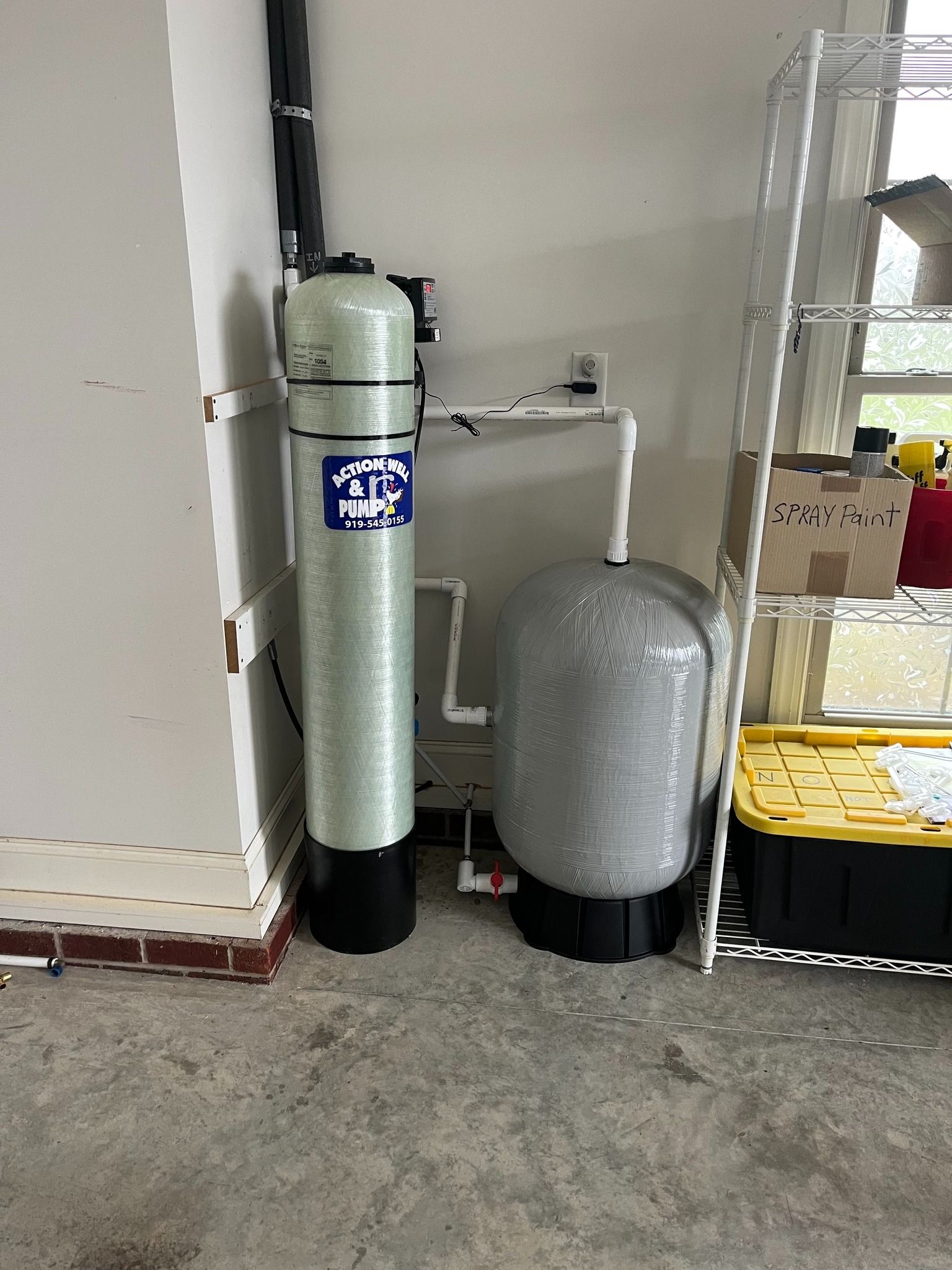 A tall, light green water filter tank next to a shorter, grey pressure tank connected by white PVC pipes in a garage.