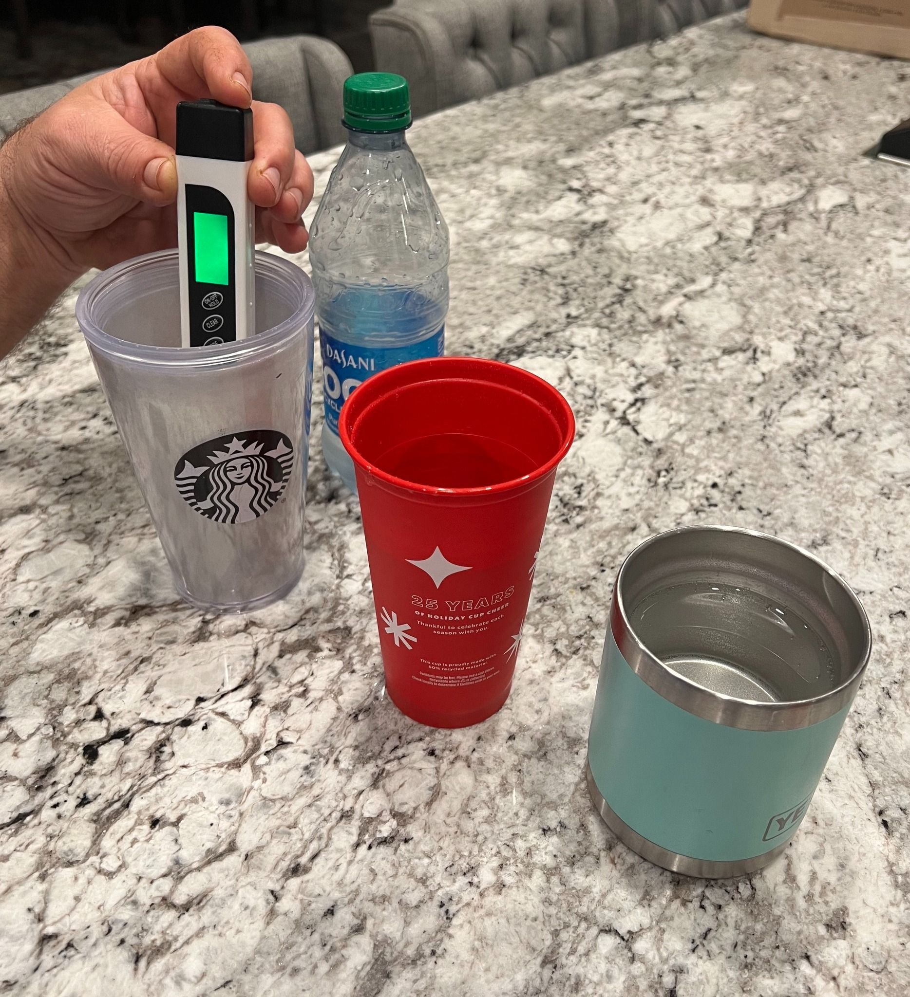 A hand holds a digital TDS meter in a clear Starbucks cup on a granite countertop with a water bottle and two other cups.