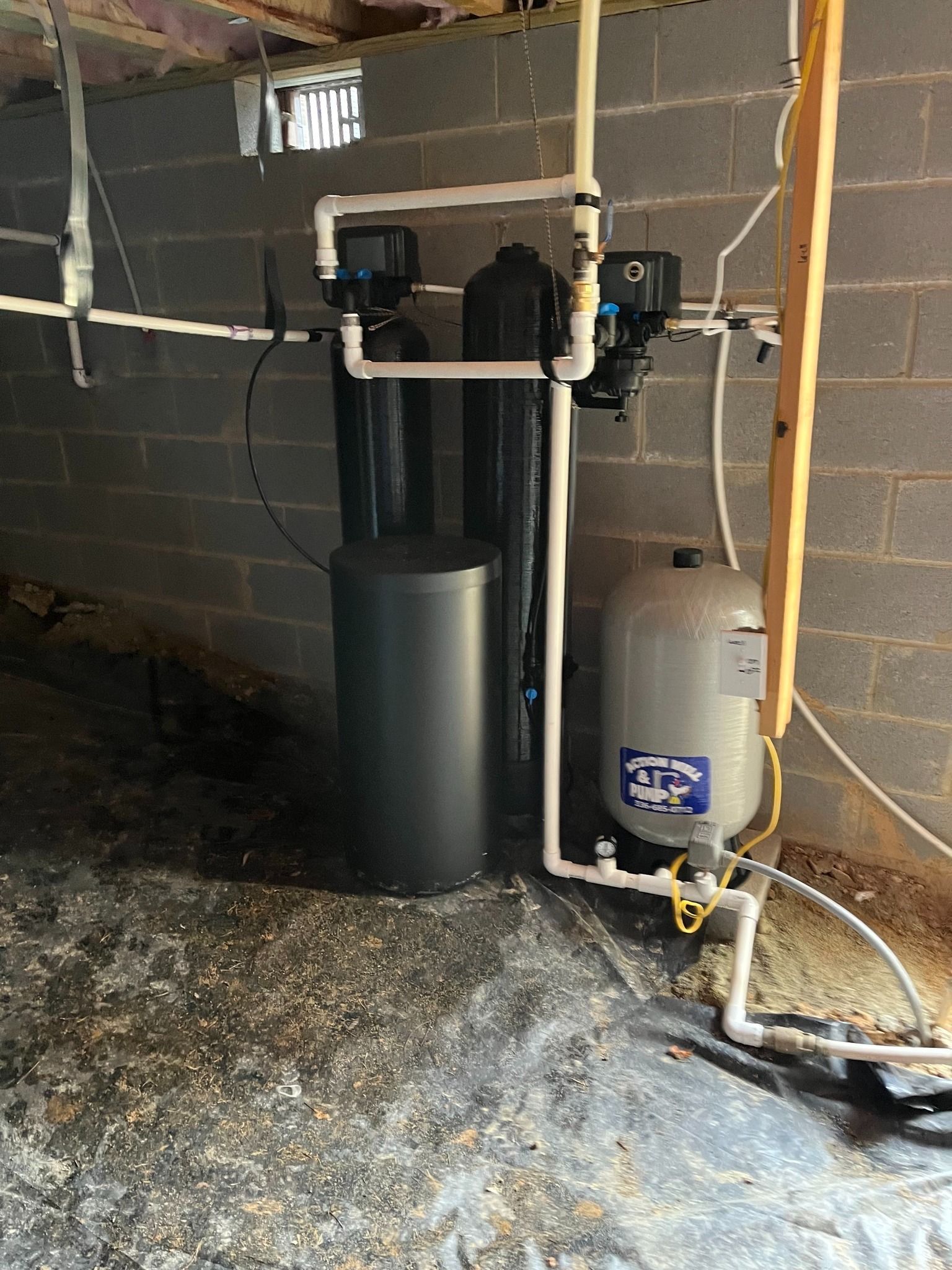 A residential water treatment system with black tanks and a grey pressure tank installed against a concrete block wall.