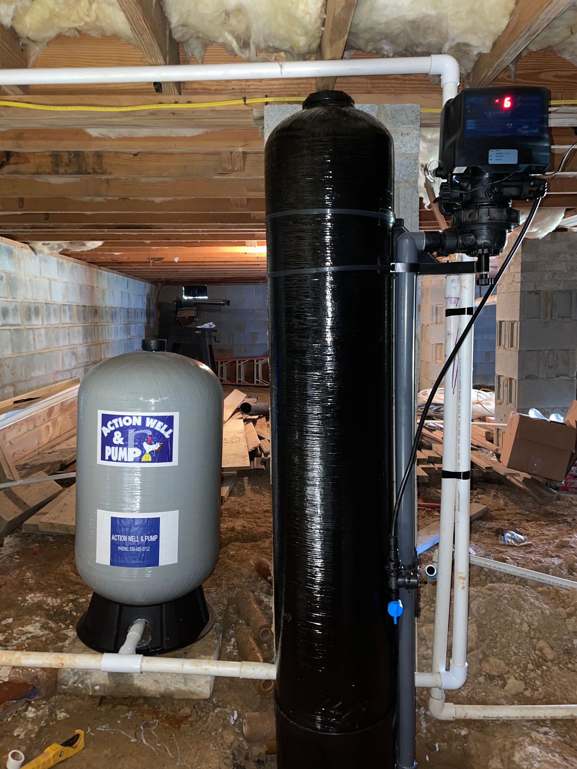 A tall black water filtration tank next to a grey pressure tank in a residential basement crawl space.